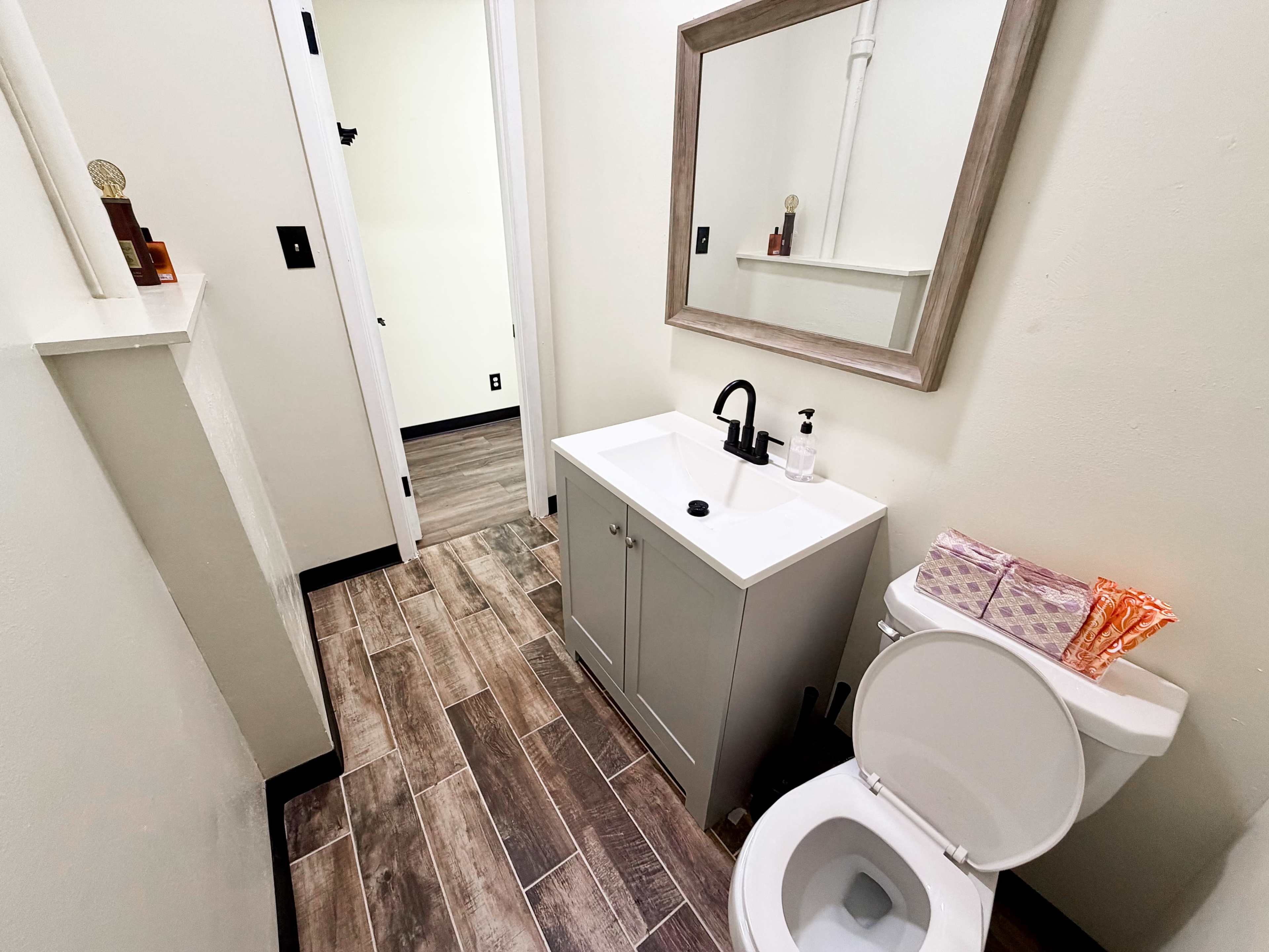 The image shows a small bathroom featuring a toilet, a sink with a vanity, and a wall-mounted mirror, with tiled flooring and neutral-colored walls.