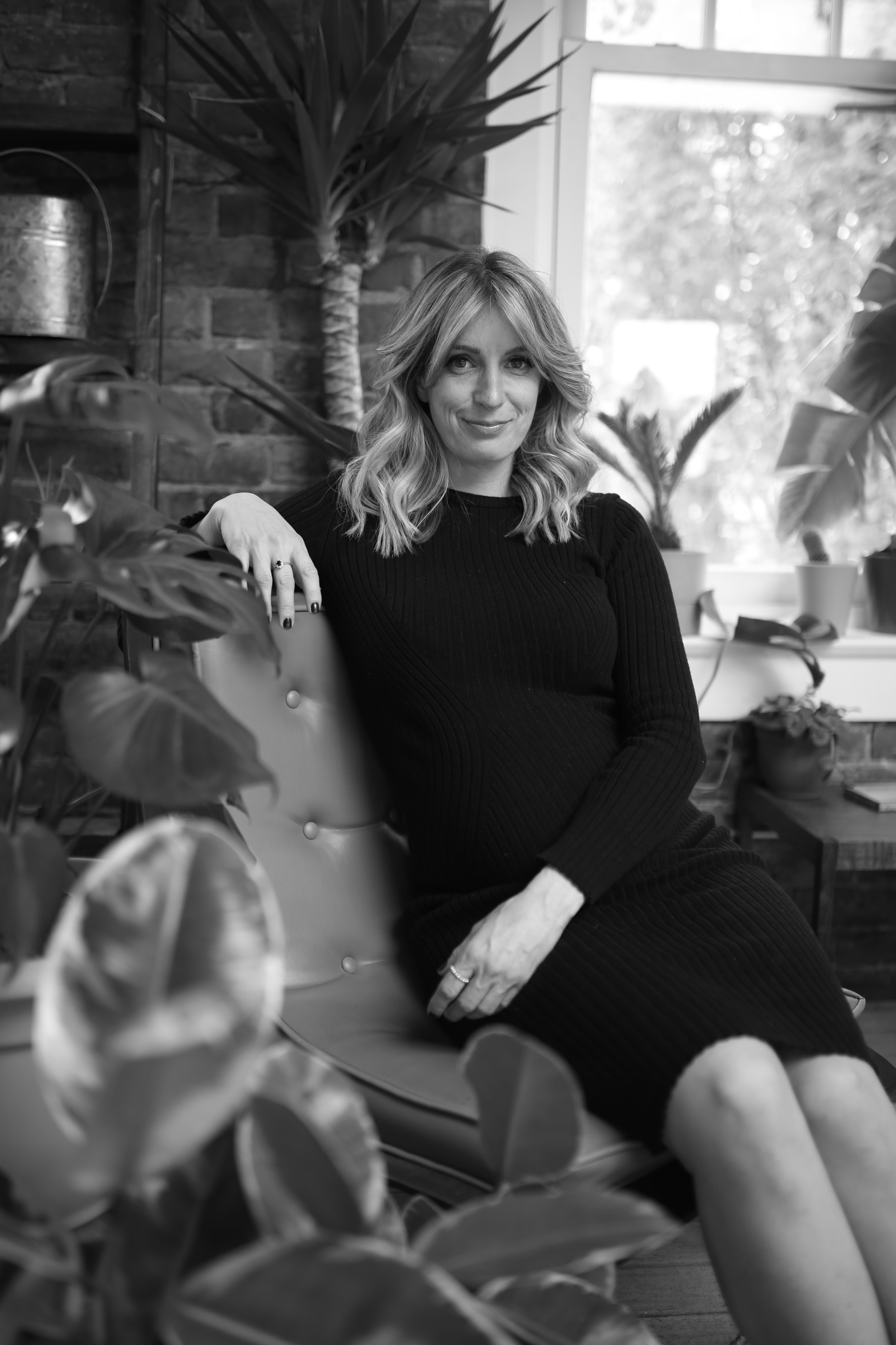 A black and white maternity photoshoot of a woman in a black dress surrounded by plants.