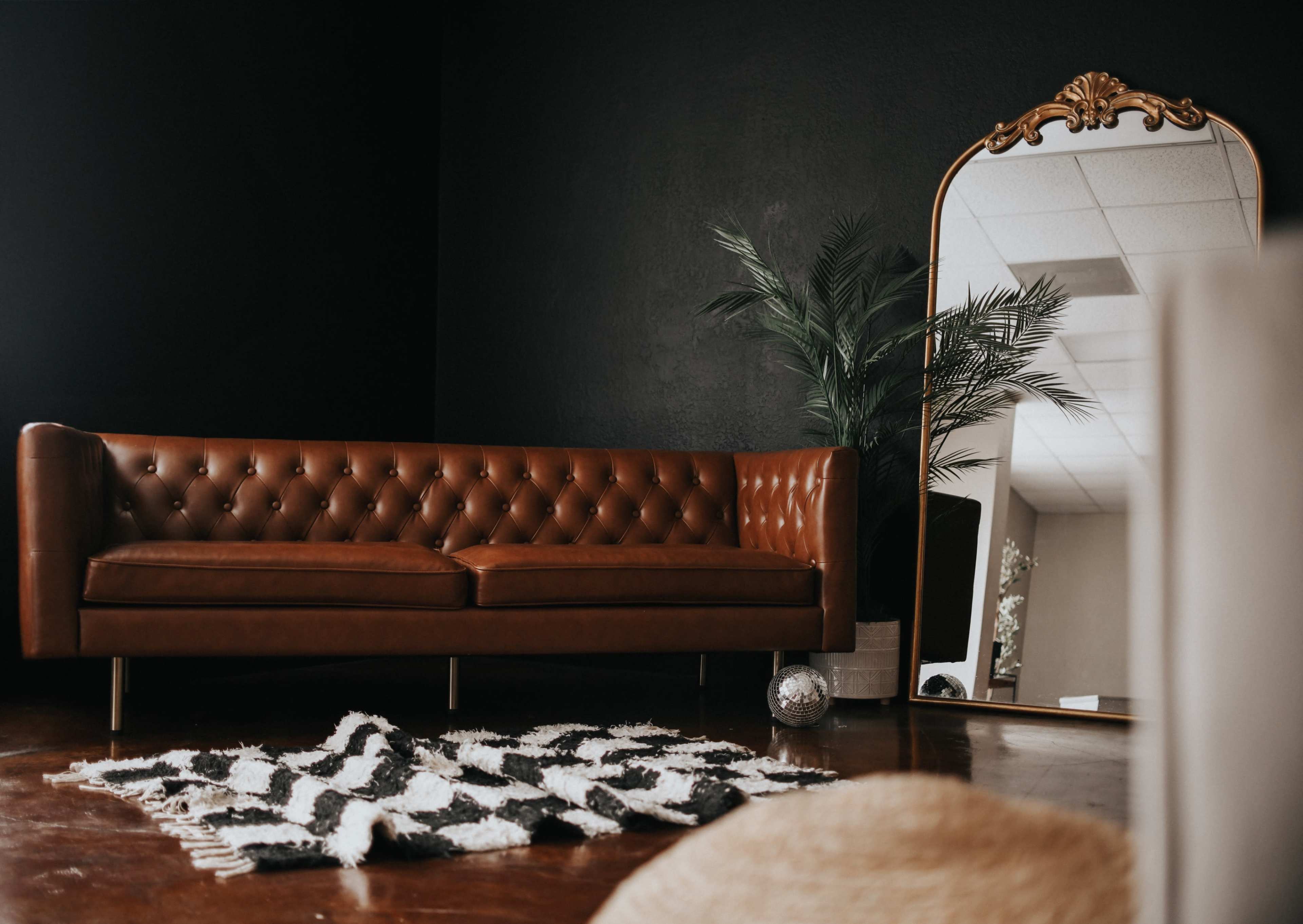 A brown leather sofa is positioned in front of a large, ornate mirror, with a striped rug and a potted plant nearby.
