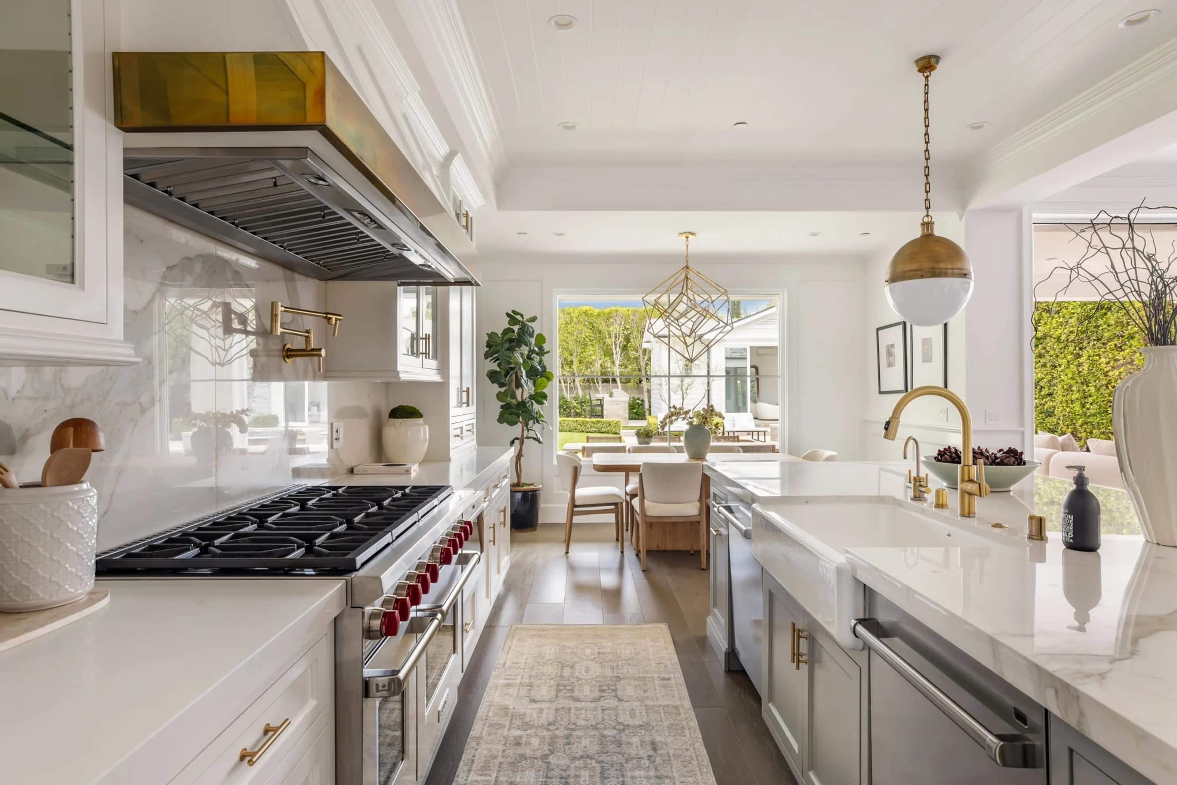The image shows a modern kitchen featuring a large gas stove, white marble countertops, and an adjacent dining area visible through a window.