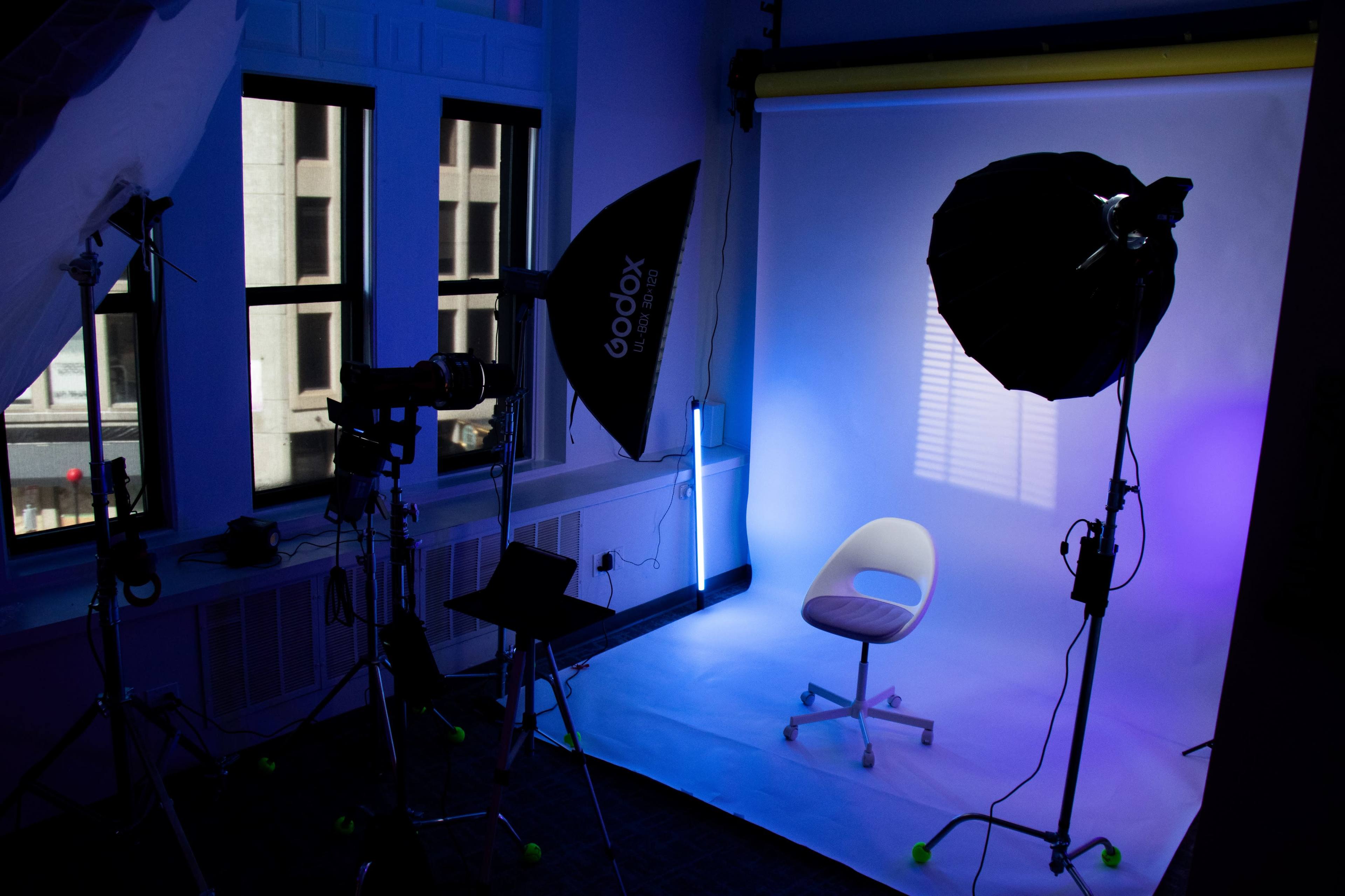 The image depicts a photography studio setup with a white backdrop, lighting equipment, and a single white chair positioned in the center.