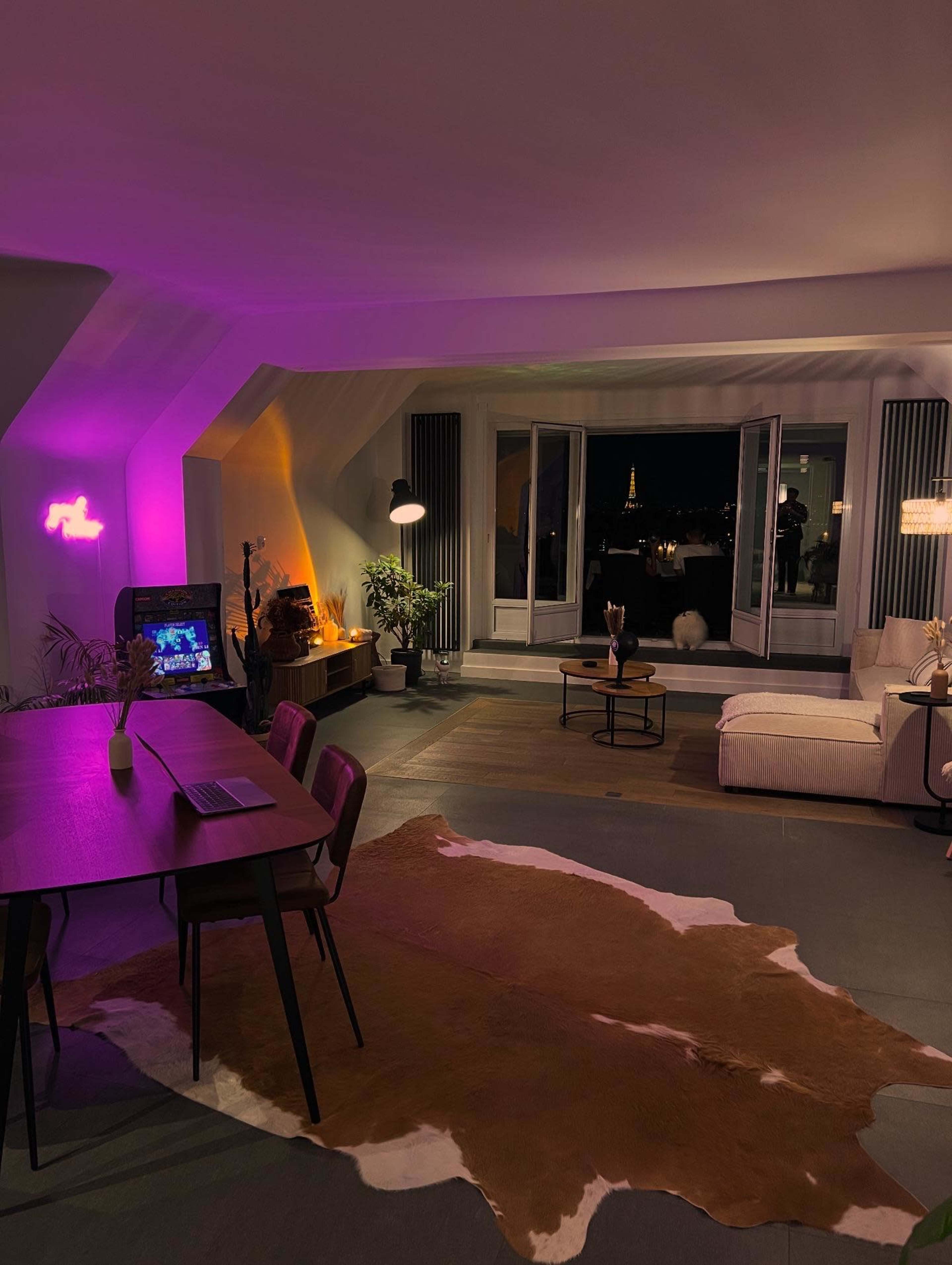 A modern living room features a dining table, a couch, and large windows overlooking the city skyline at night, illuminated by soft purple lights.