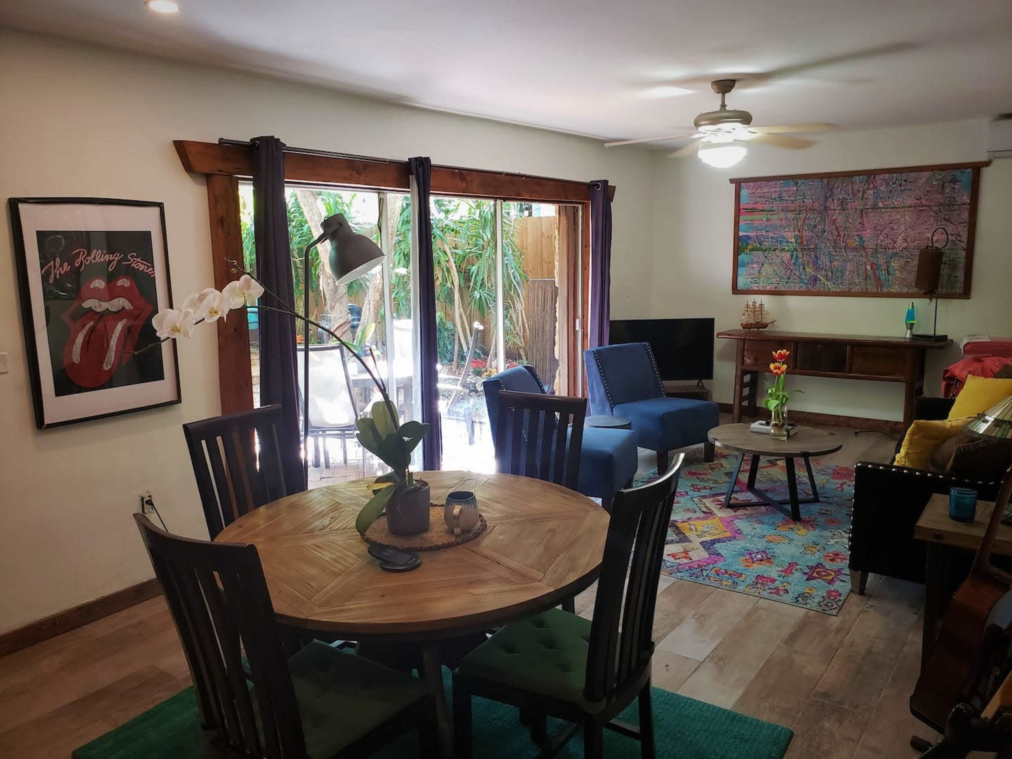 A living room features a round dining table with chairs, a green area rug, and large glass doors opening to an outdoor space with greenery.