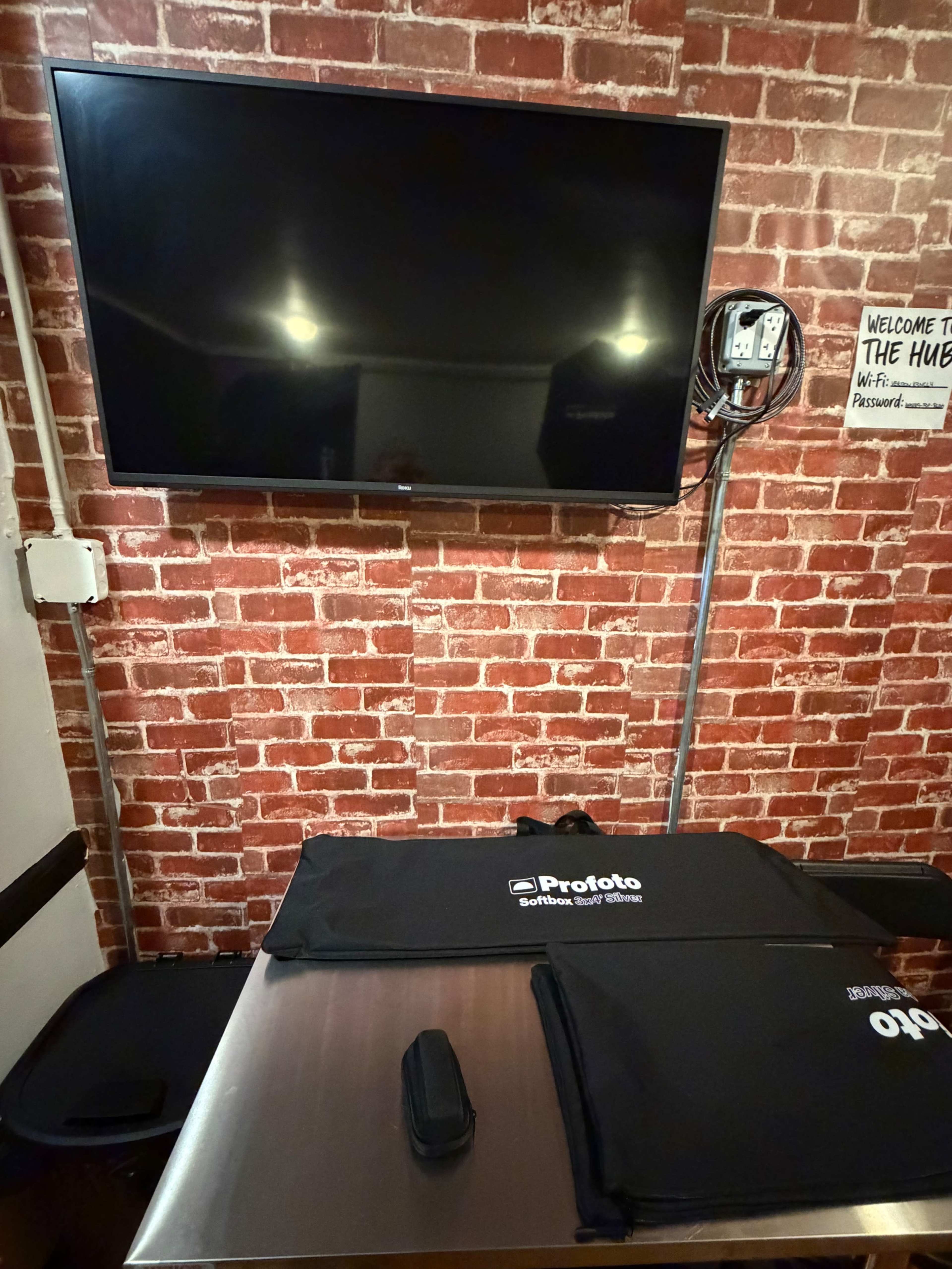 The image shows a metal table with black bags labeled "Profoto" on it, a wall made of exposed bricks in the background, and a large flat-screen TV mounted above the table.
