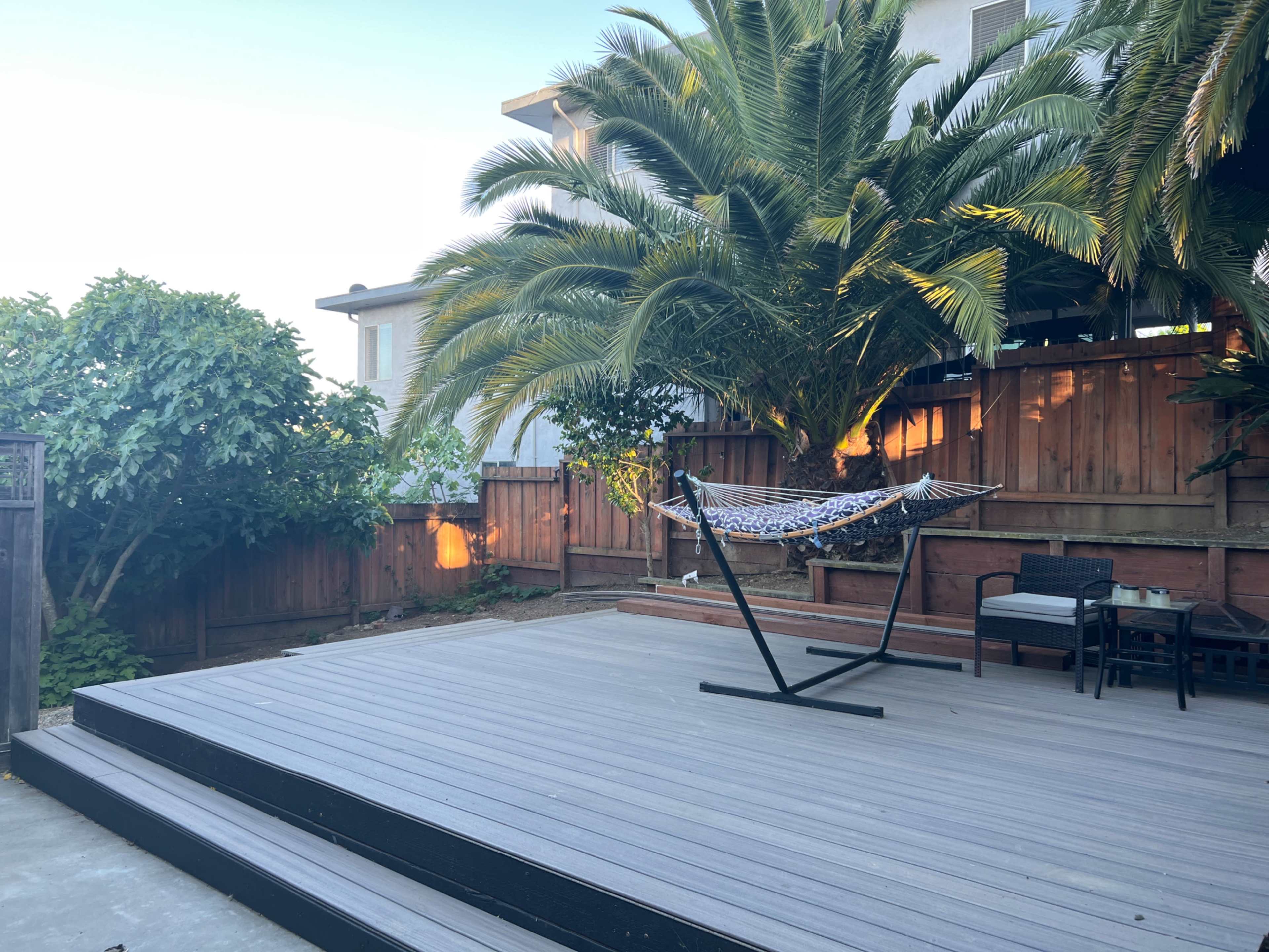 A wooden deck features a hammock hanging between palm trees, with a seating area in the background.