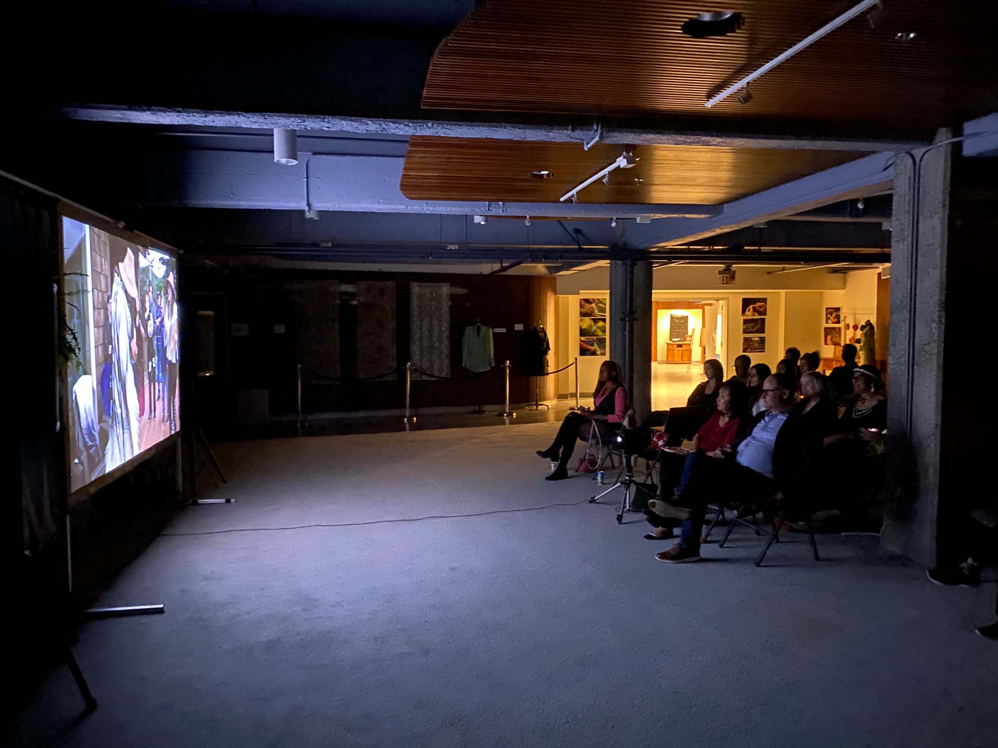 A group of people is seated in chairs facing a large screen displaying a video in a dimly lit room.