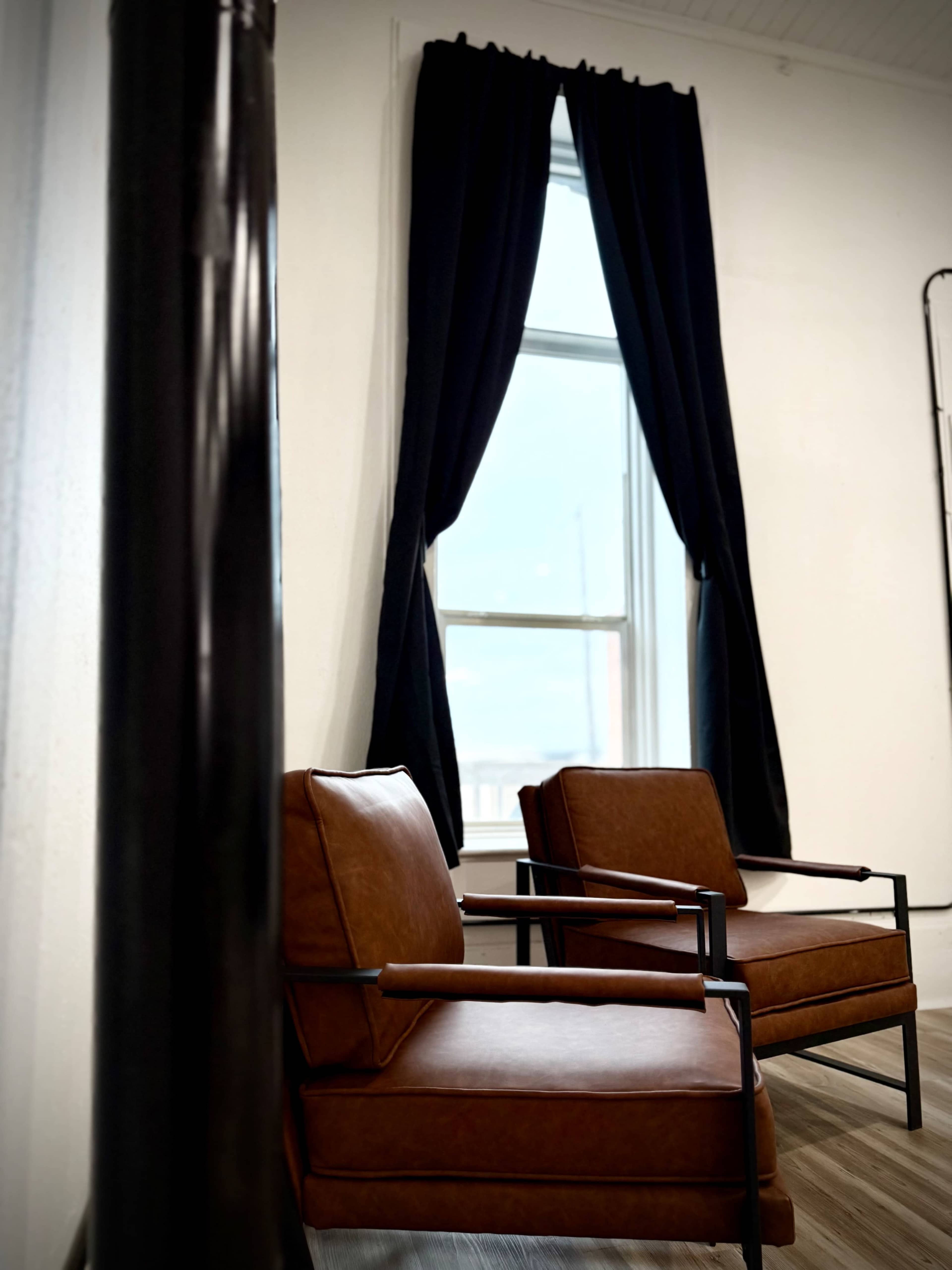 Two brown leather chairs are positioned beside a large window covered with black curtains in a modern, minimalistic interior space.