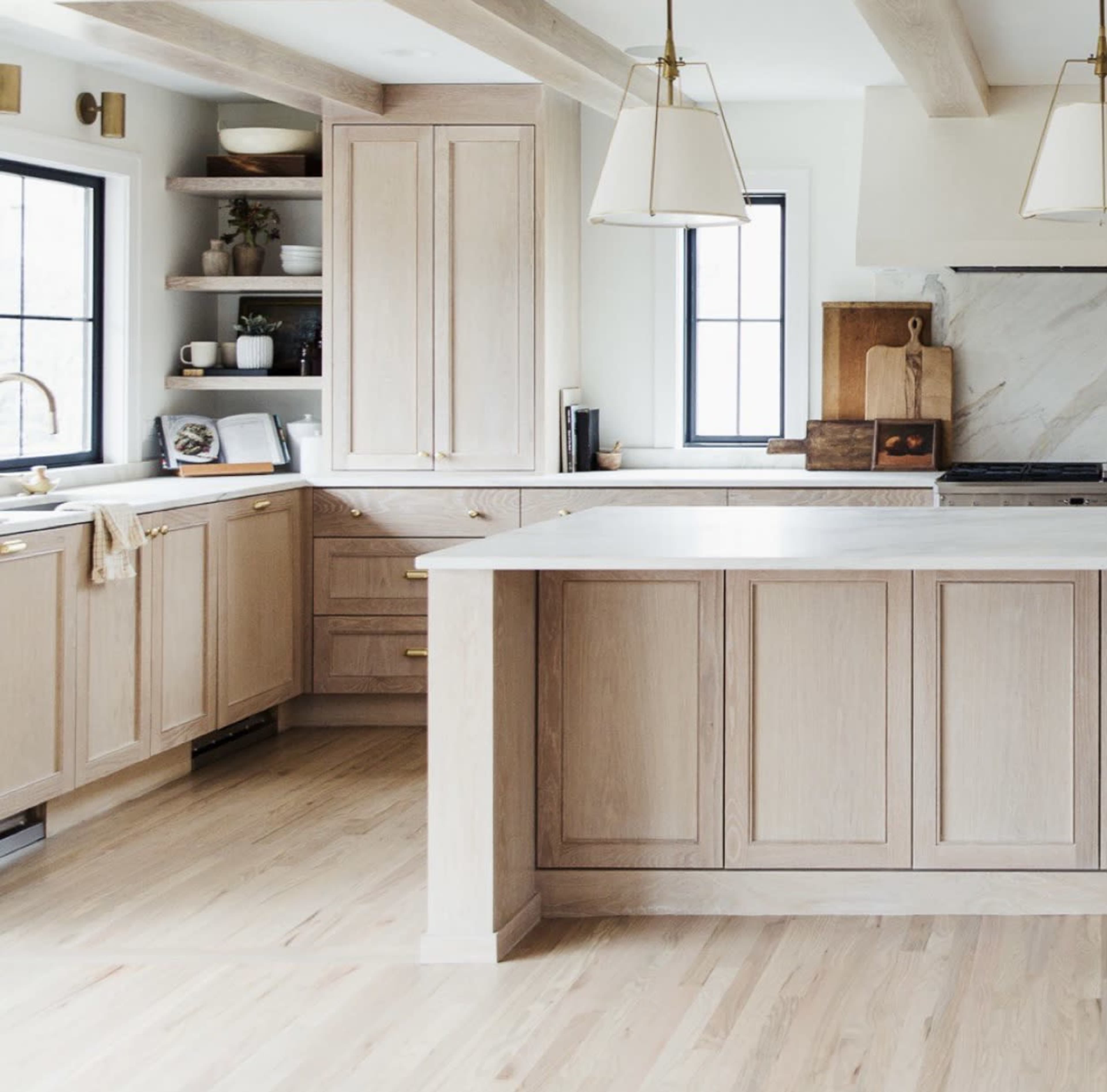 The image shows a modern kitchen featuring light wooden cabinetry, a center island, and large windows that allow natural light to enter.