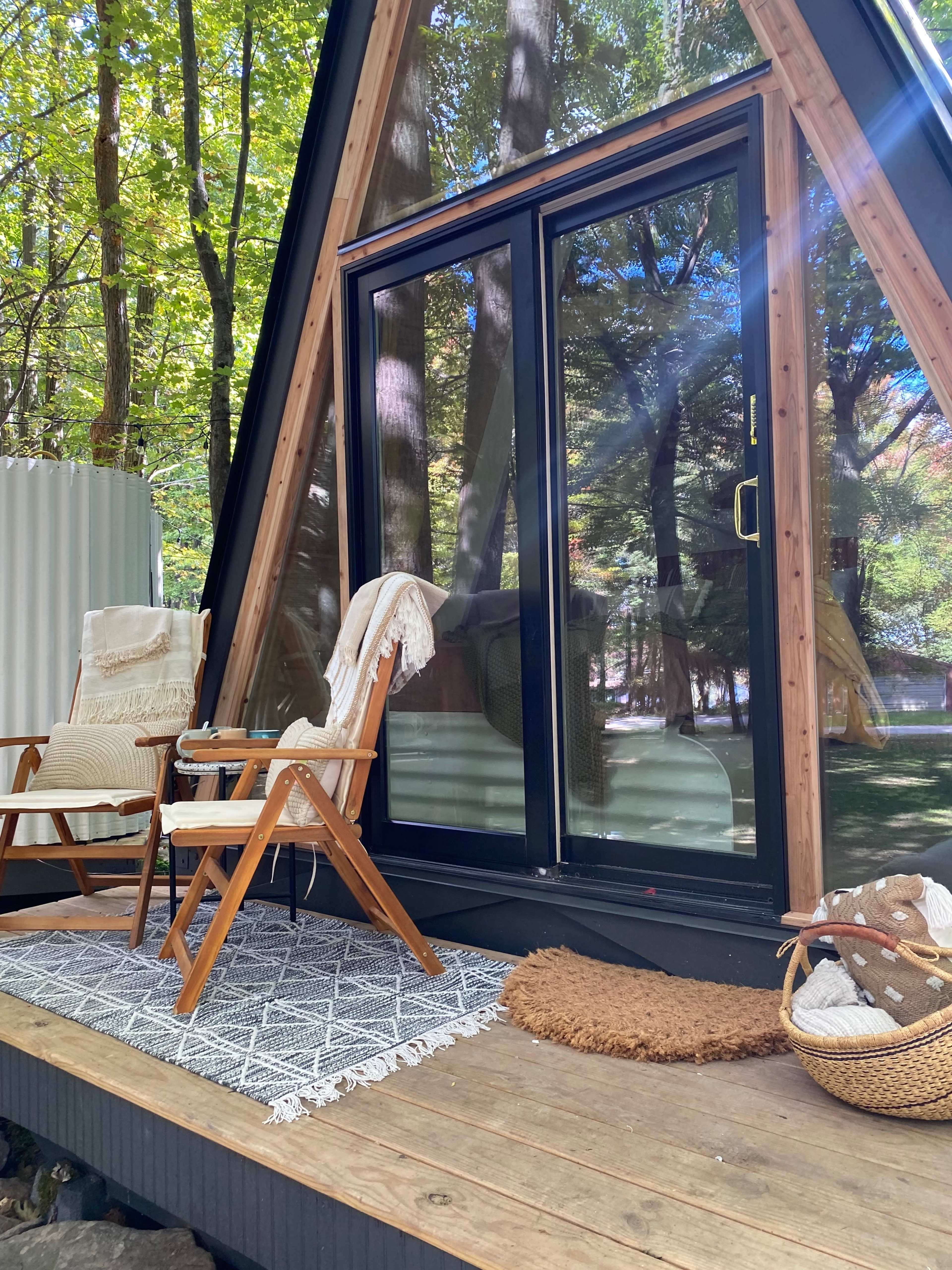 A cozy A-frame cabin porch with two wooden chairs, a patterned rug, and a woven basket, surrounded by trees.
