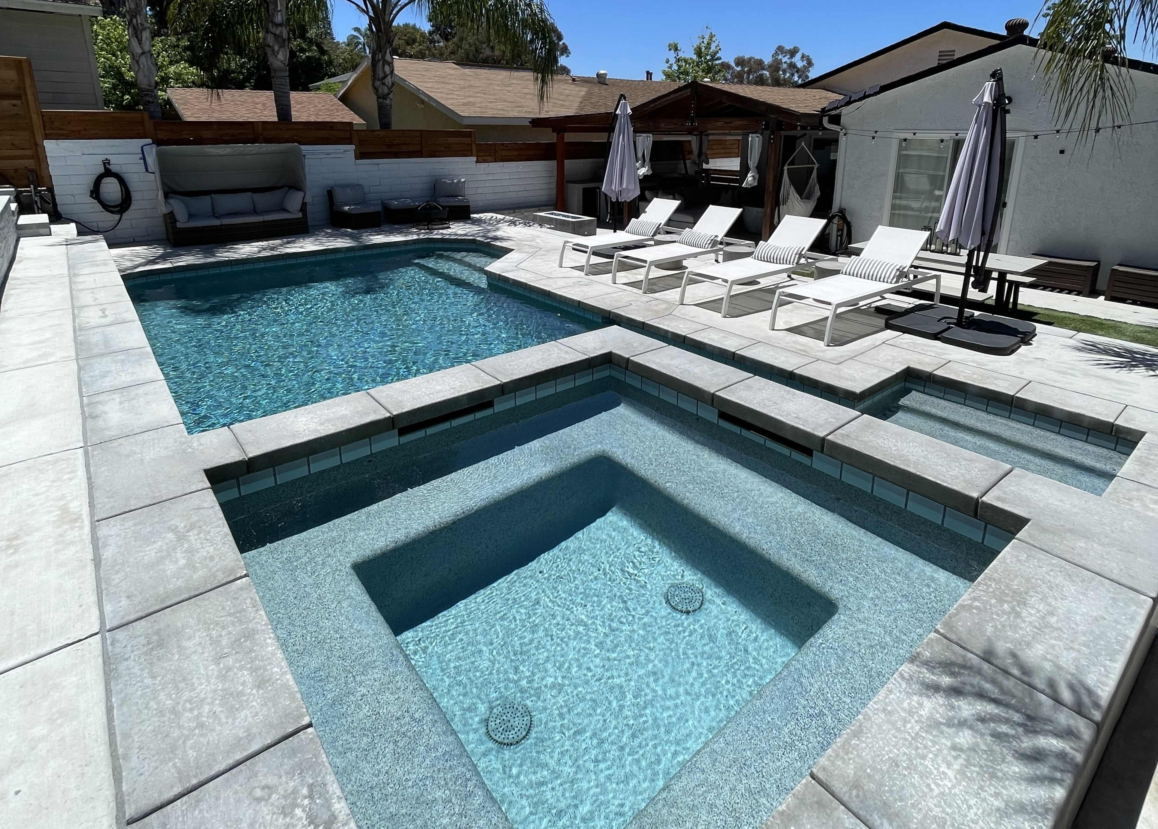 A clear swimming pool with a spa area is surrounded by lounge chairs and shaded umbrellas in a residential backyard.