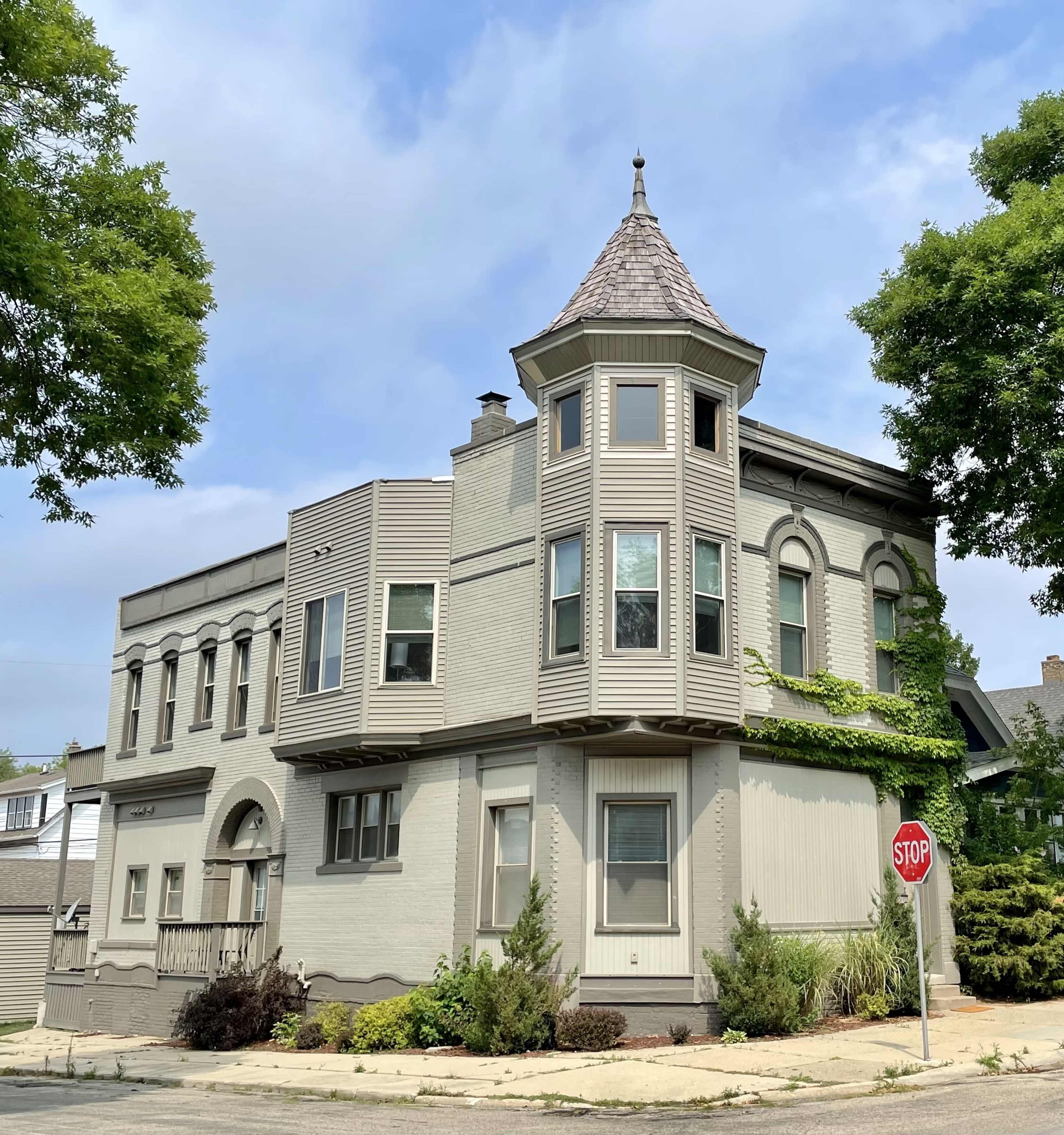 A gray, multi-story building with a distinctive turret and large windows is located at the corner of a quiet street, featuring landscaped greenery and a stop sign nearby.