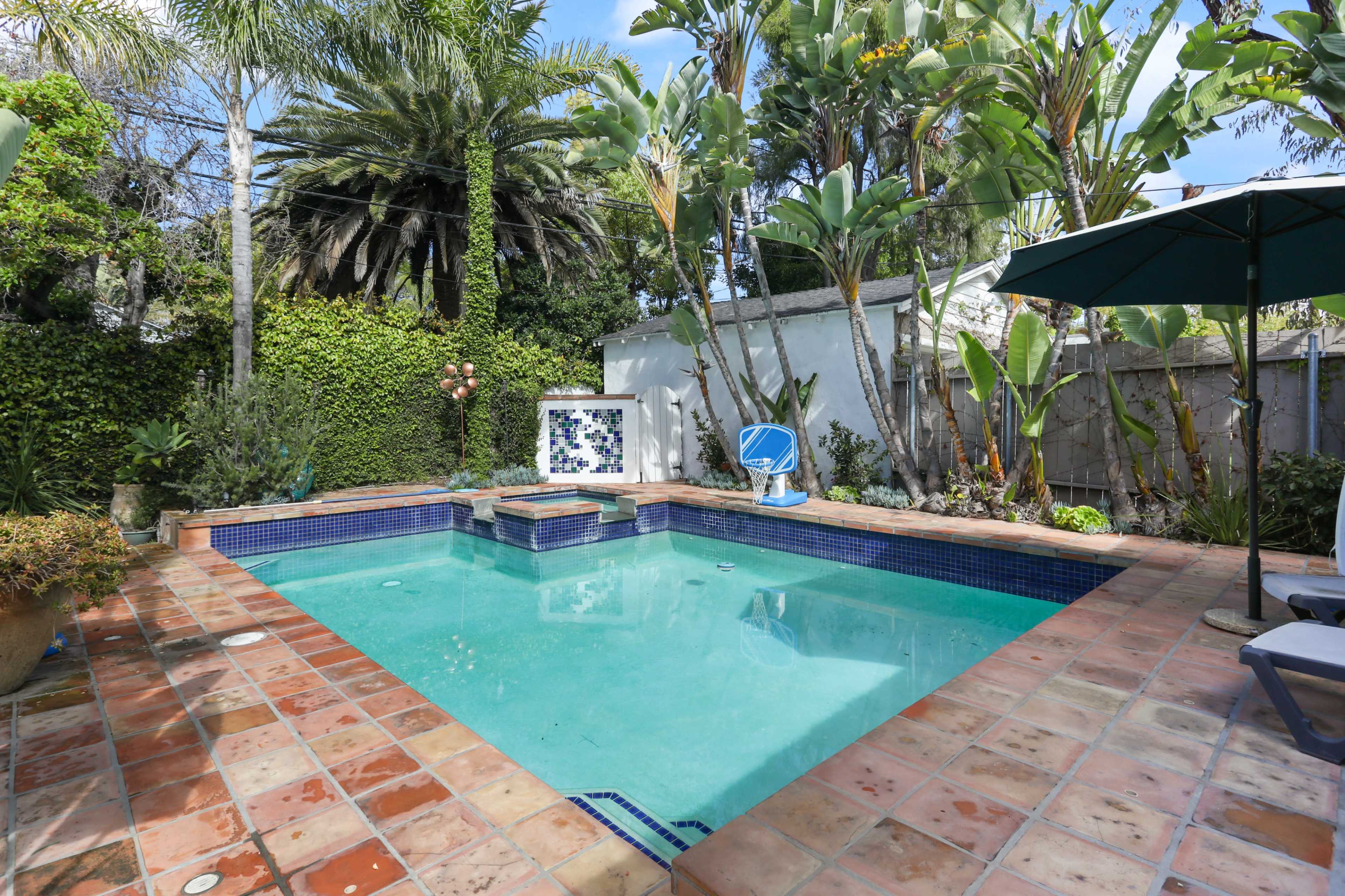 A backyard pool surrounded by lush greenery, featuring a tiled patio and a lounge area with an umbrella.
