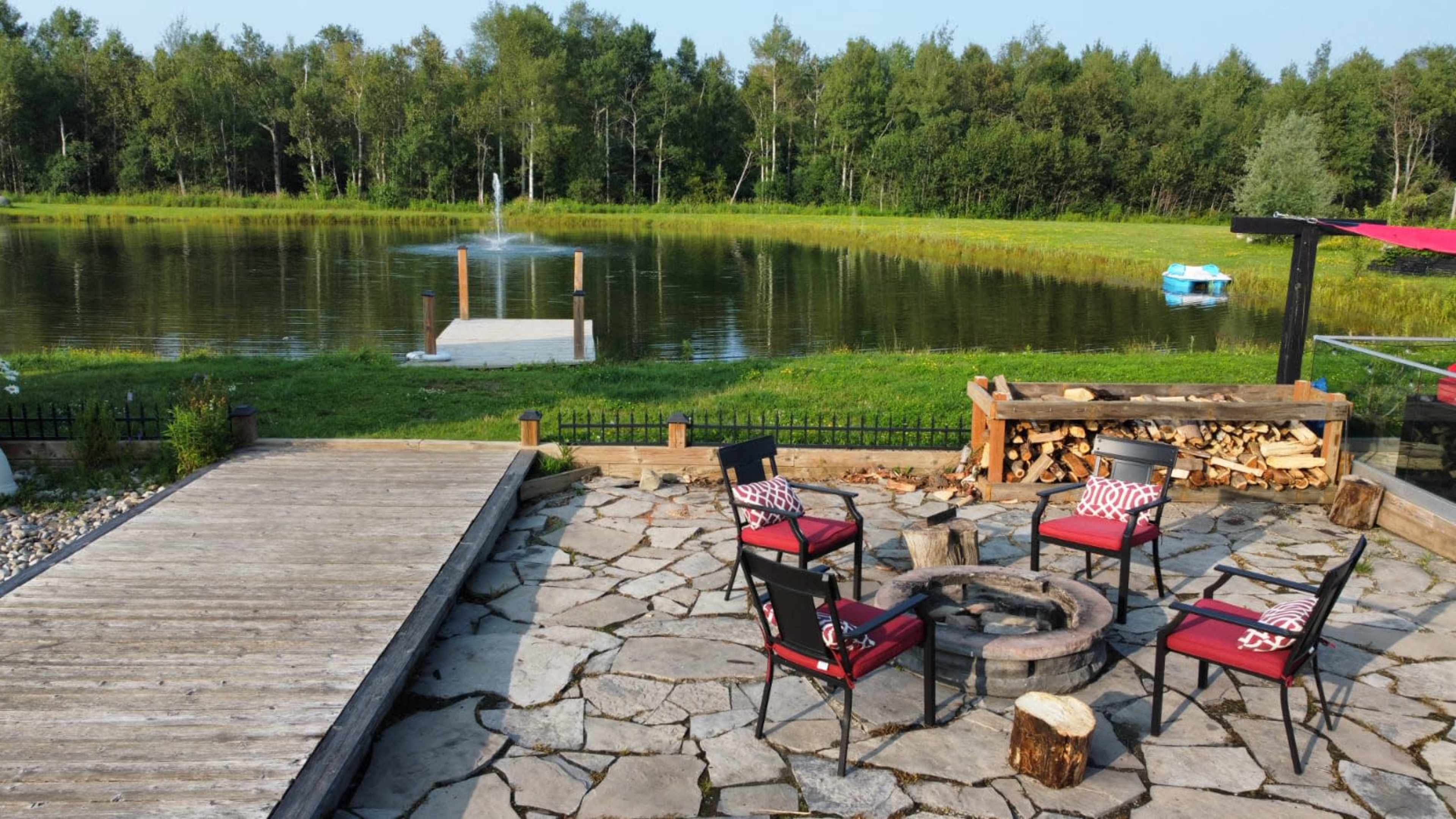 A stone patio with four chairs arranged around a fire pit, situated beside a pond with a fountain and a small dock.