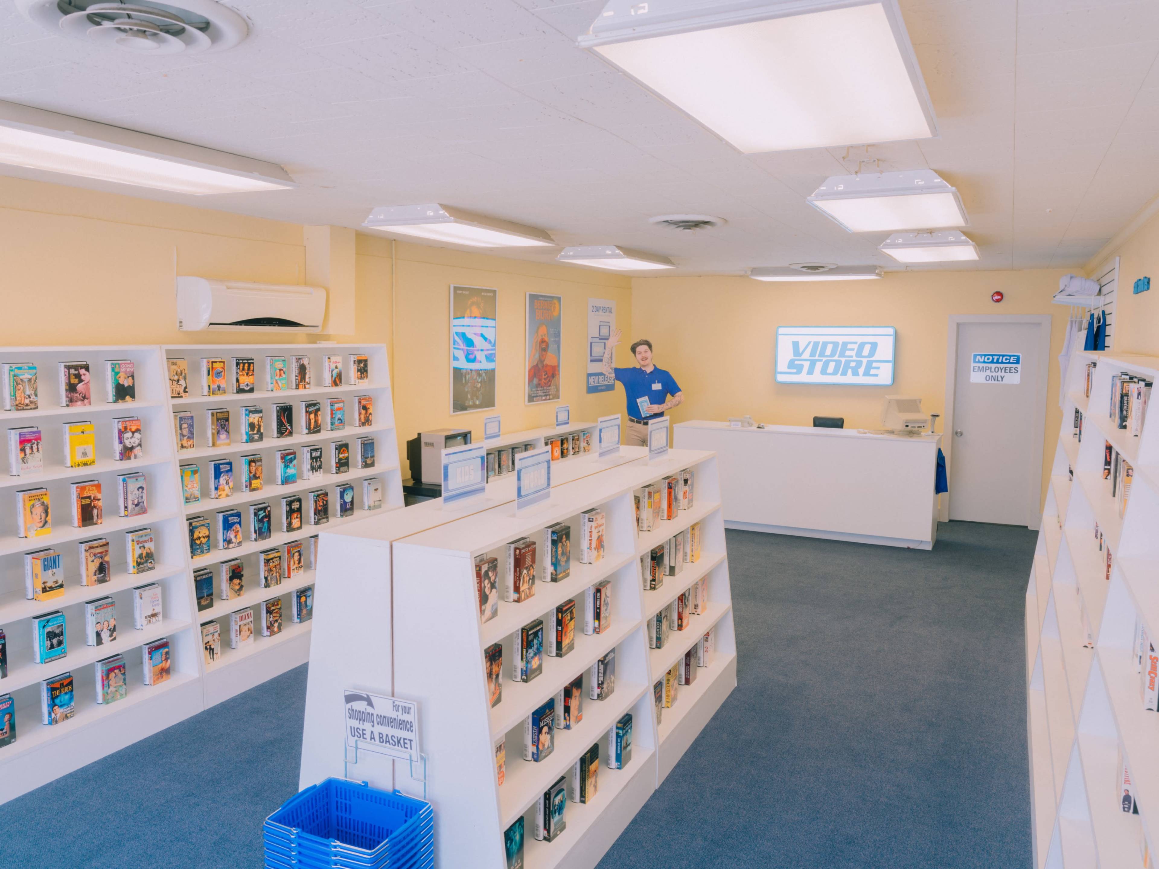 The interior of a video rental store features rows of shelves filled with DVDs and a counter with a staff member standing behind it.