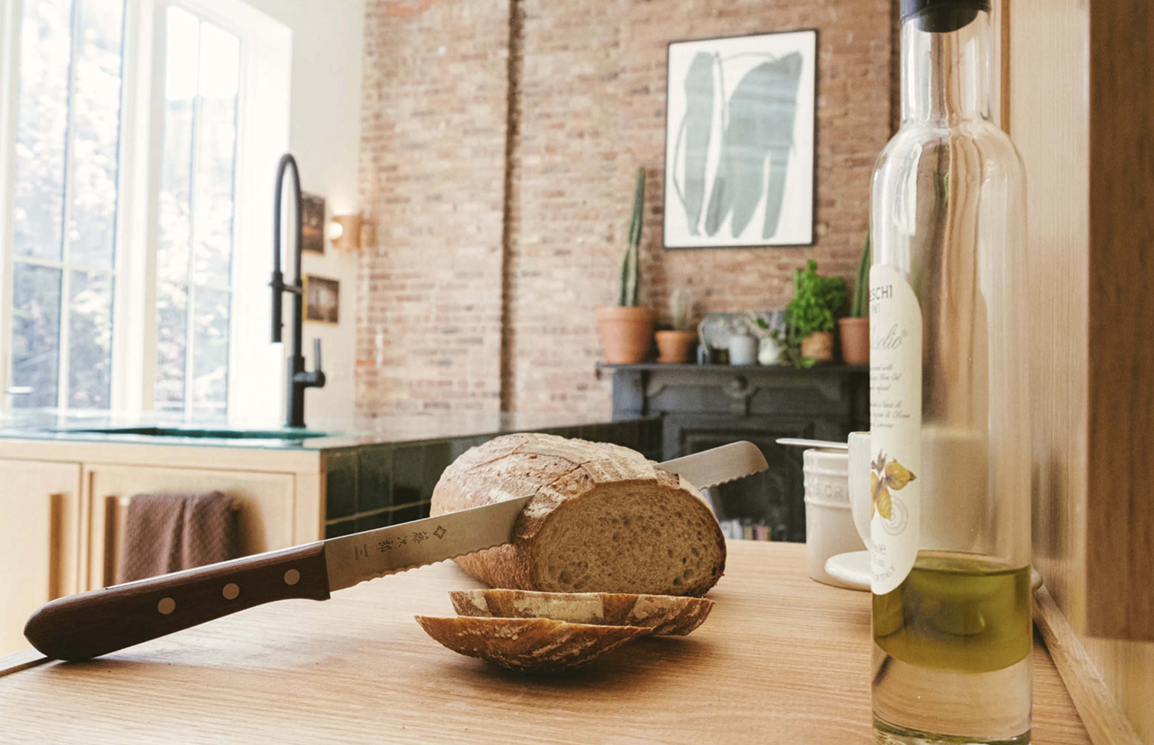 A wooden cutting board with a sliced loaf of bread, a knife, and a bottle of olive oil is set in a kitchen featuring brick walls and plants.