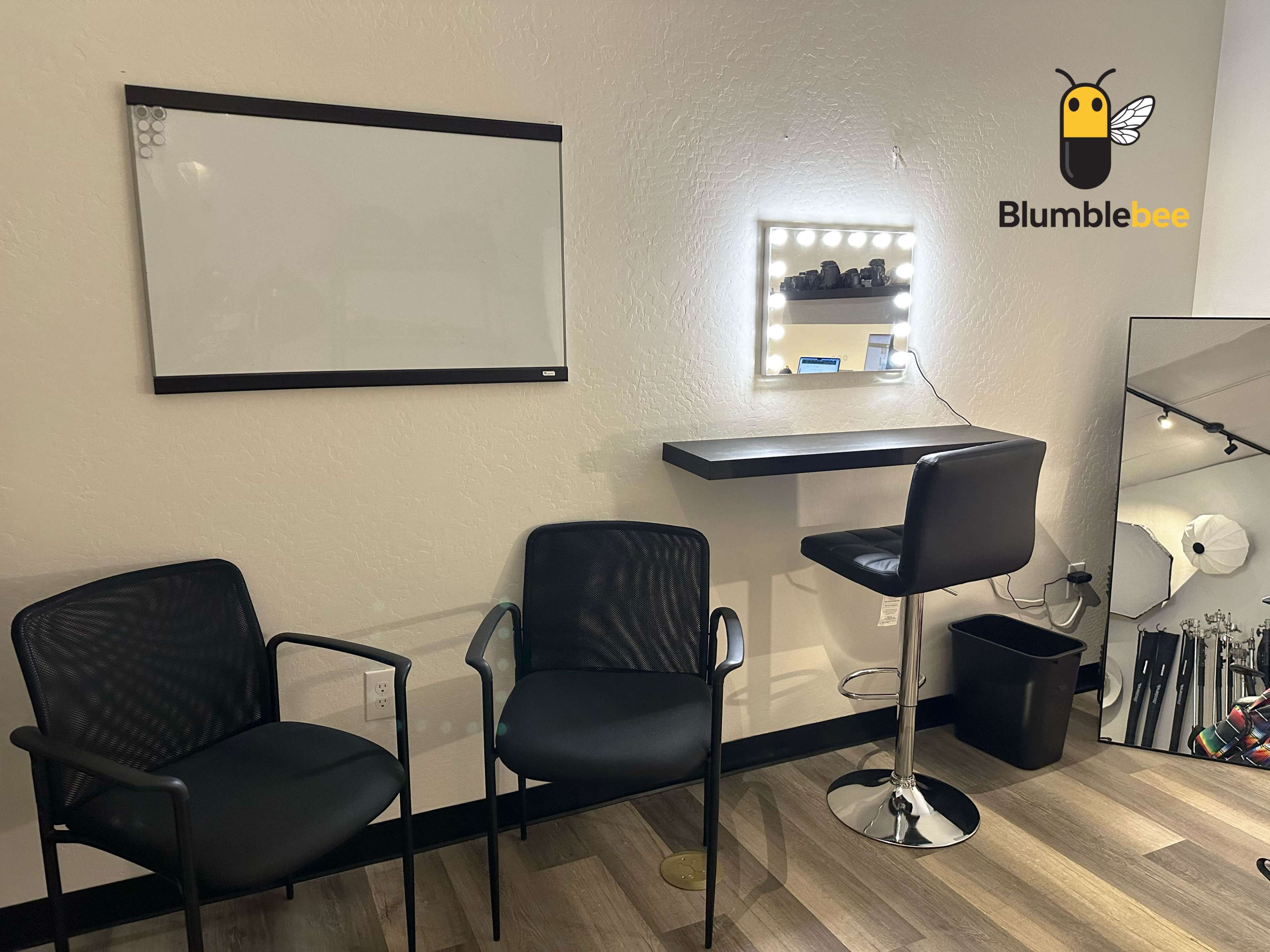 The image shows a small room with two black chairs, a mounted mirror, a lighted vanity table, and a whiteboard on the wall.