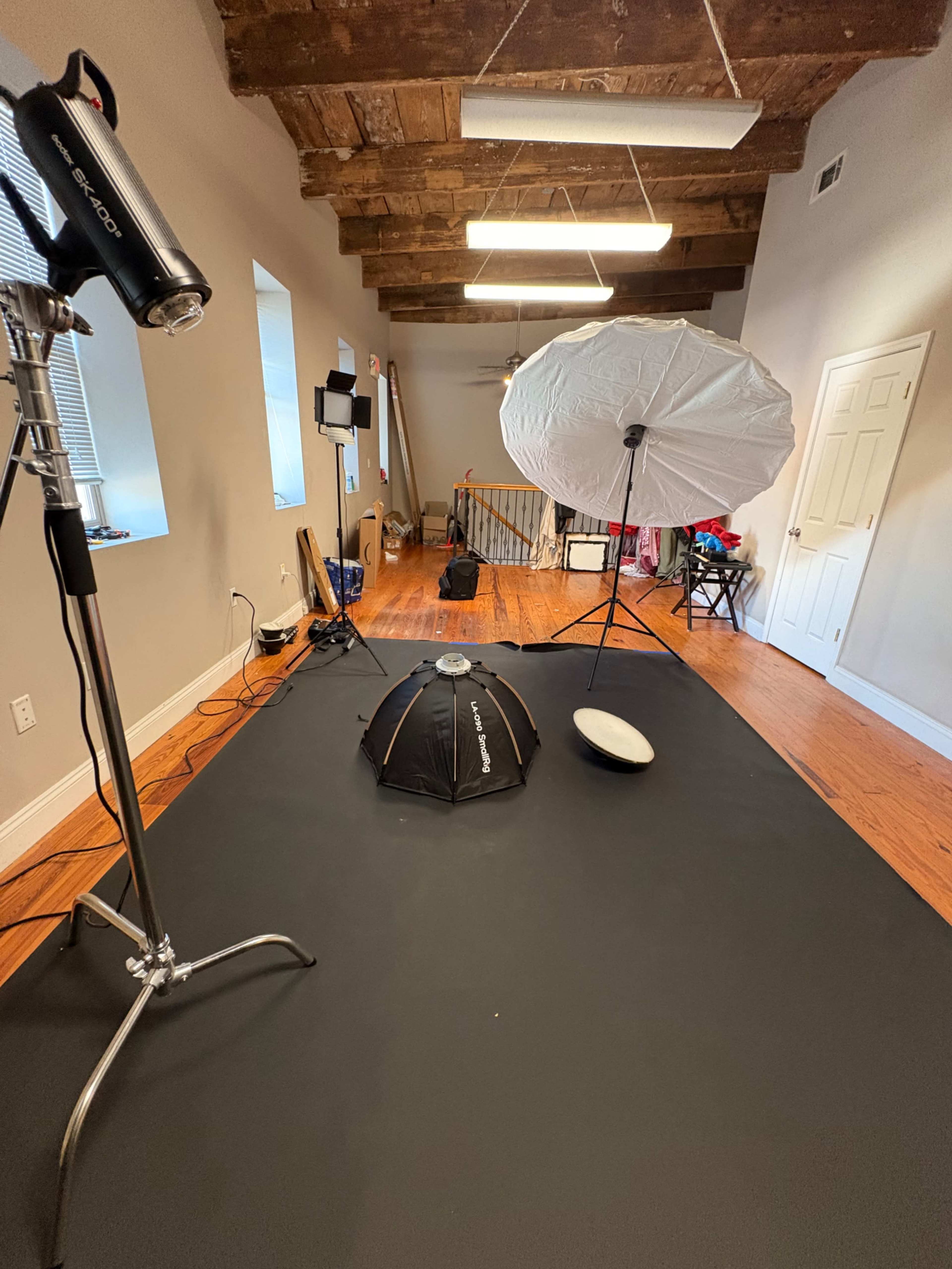 The image shows a photography setup in a room with wooden beams and a black floor, featuring various lighting equipment and umbrellas arranged on a mat.