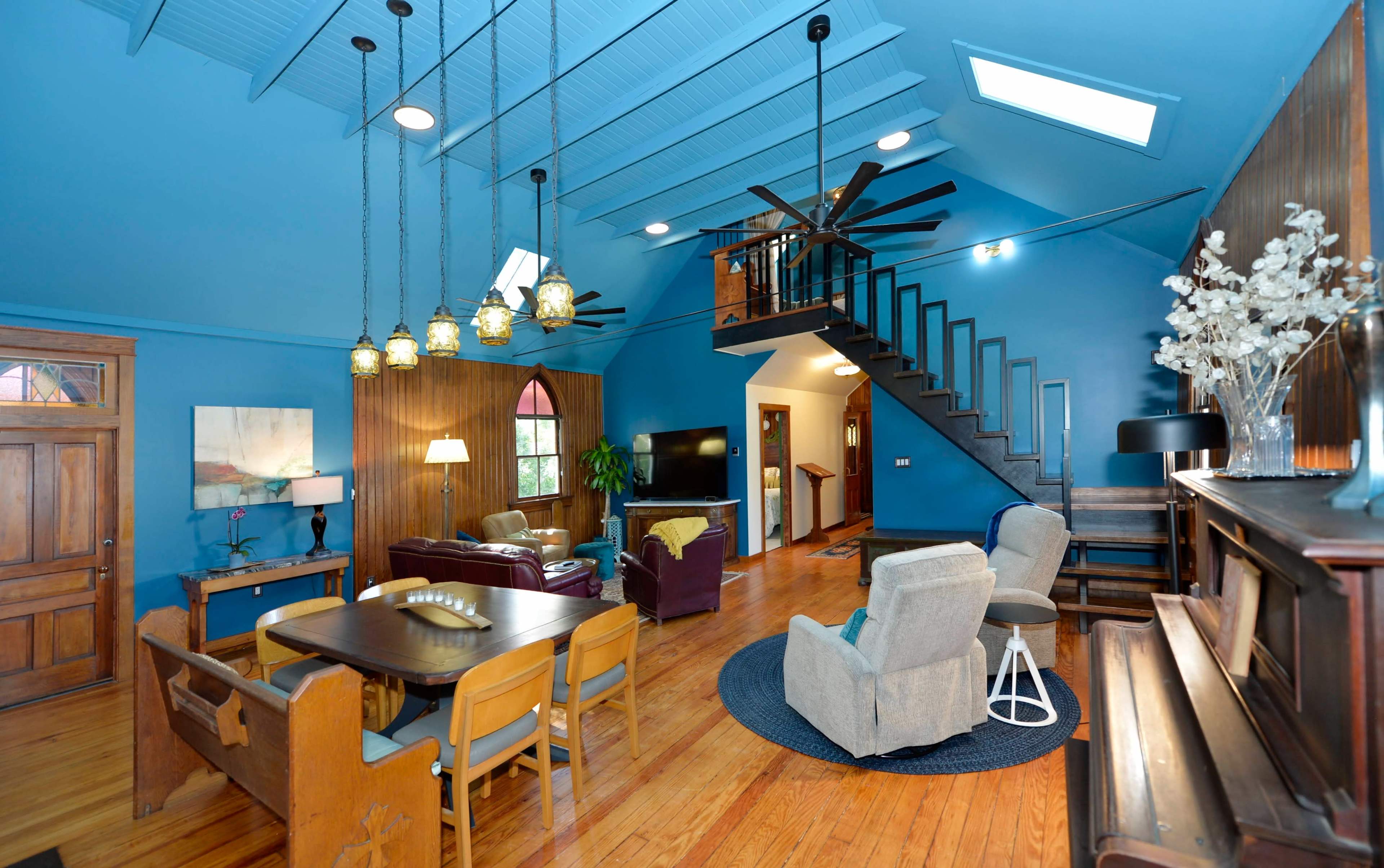 The image shows a brightly colored living area with a wooden floor, featuring a dining table, comfortable seating arrangements, large windows, and a staircase leading to an upper level.