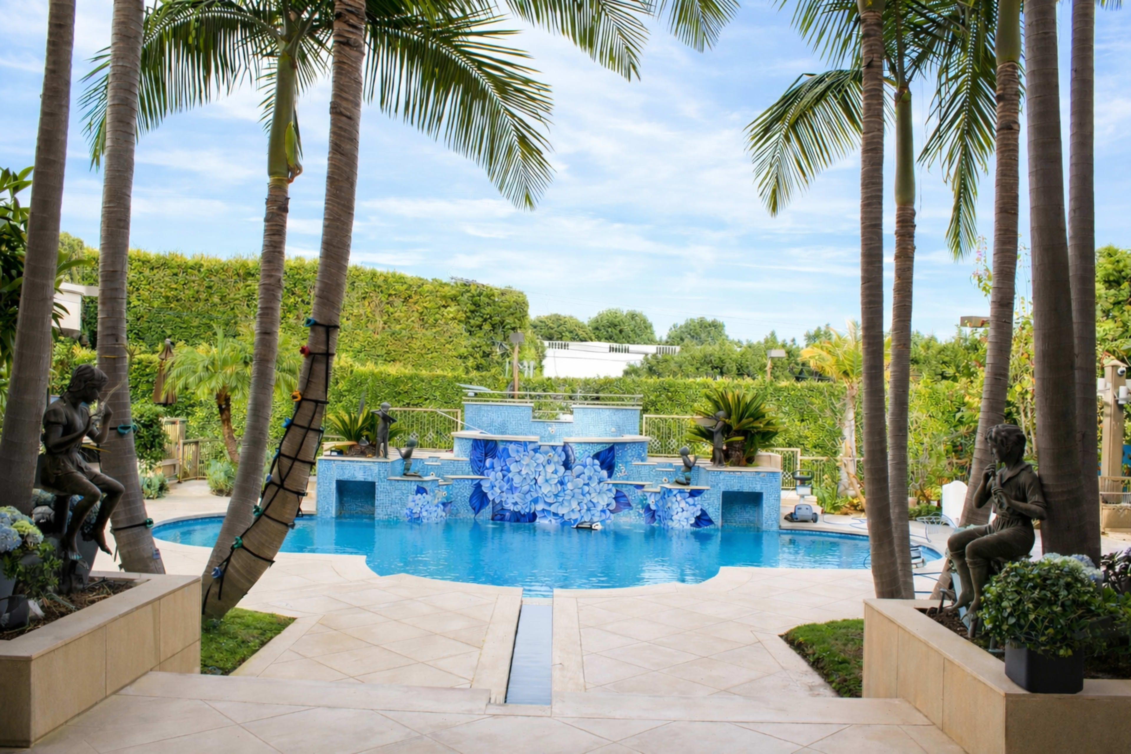 A landscaped backyard features a swimming pool with a decorative waterfall and palm trees framing the view.