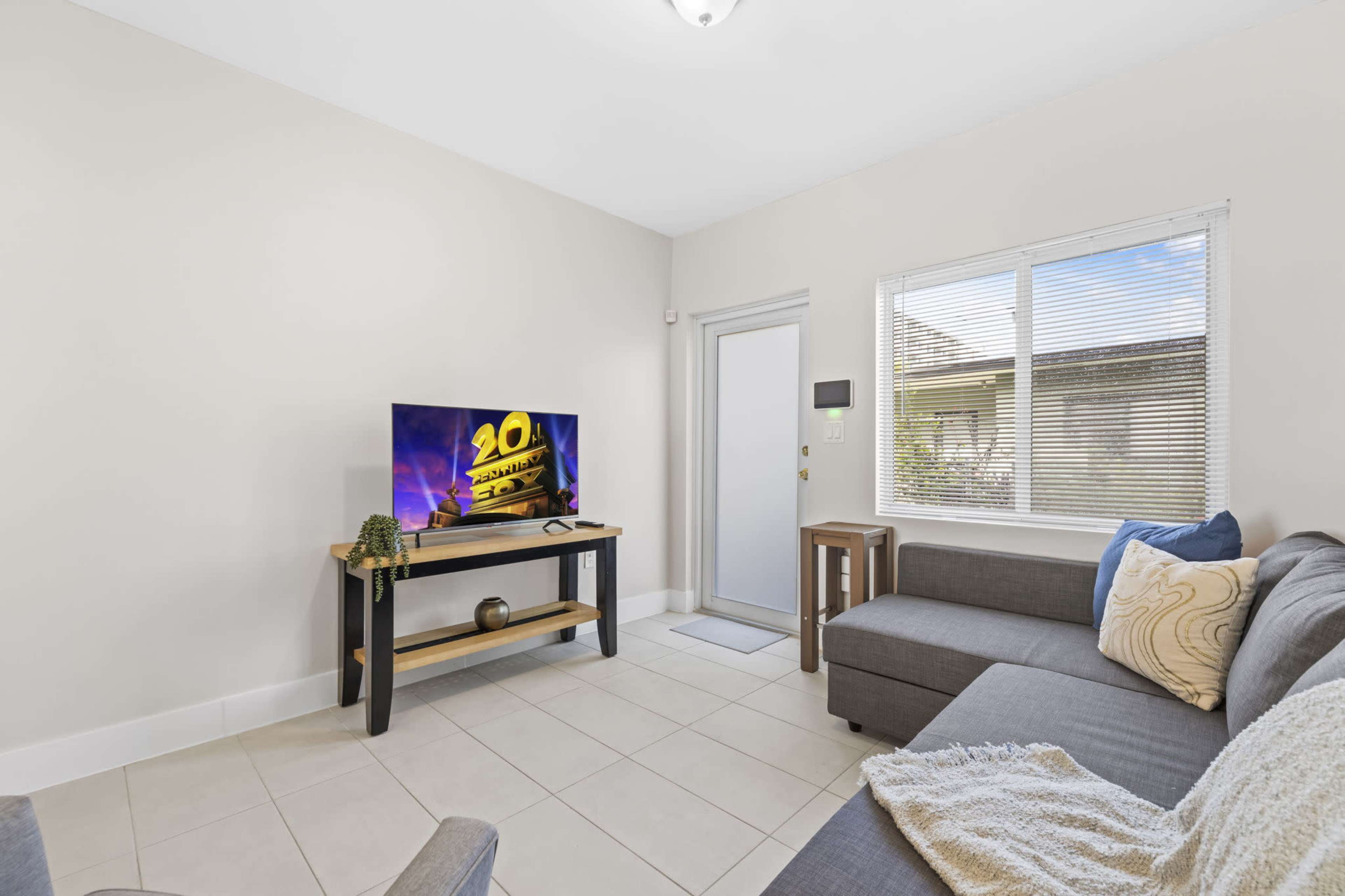 A living room features a gray sectional sofa, a television on a wooden stand, and a door leading outside, with light streaming through a window.