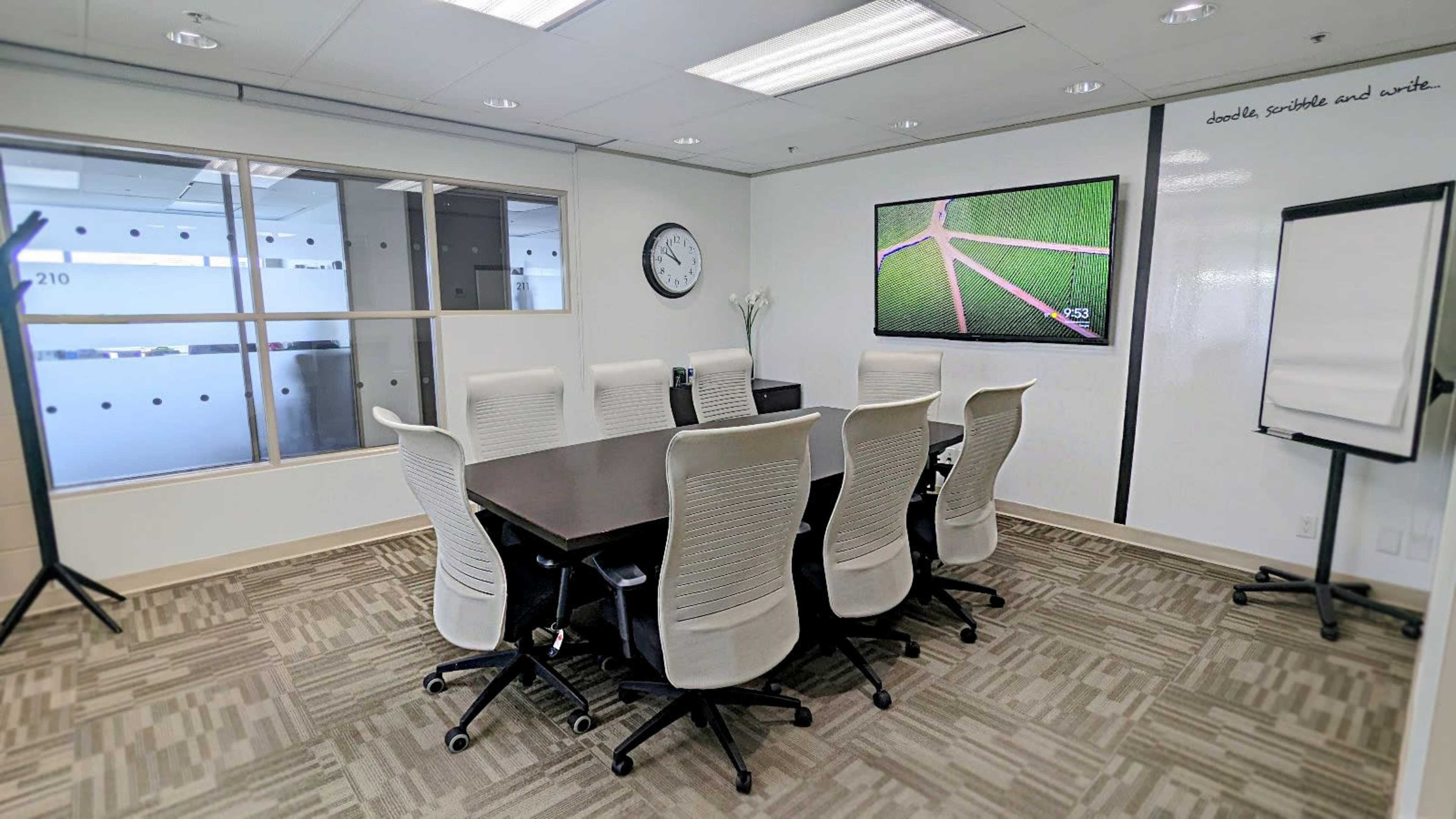 A modern conference room features a large table surrounded by seven white chairs, a wall-mounted screen displaying green fields, and a clock on the wall.