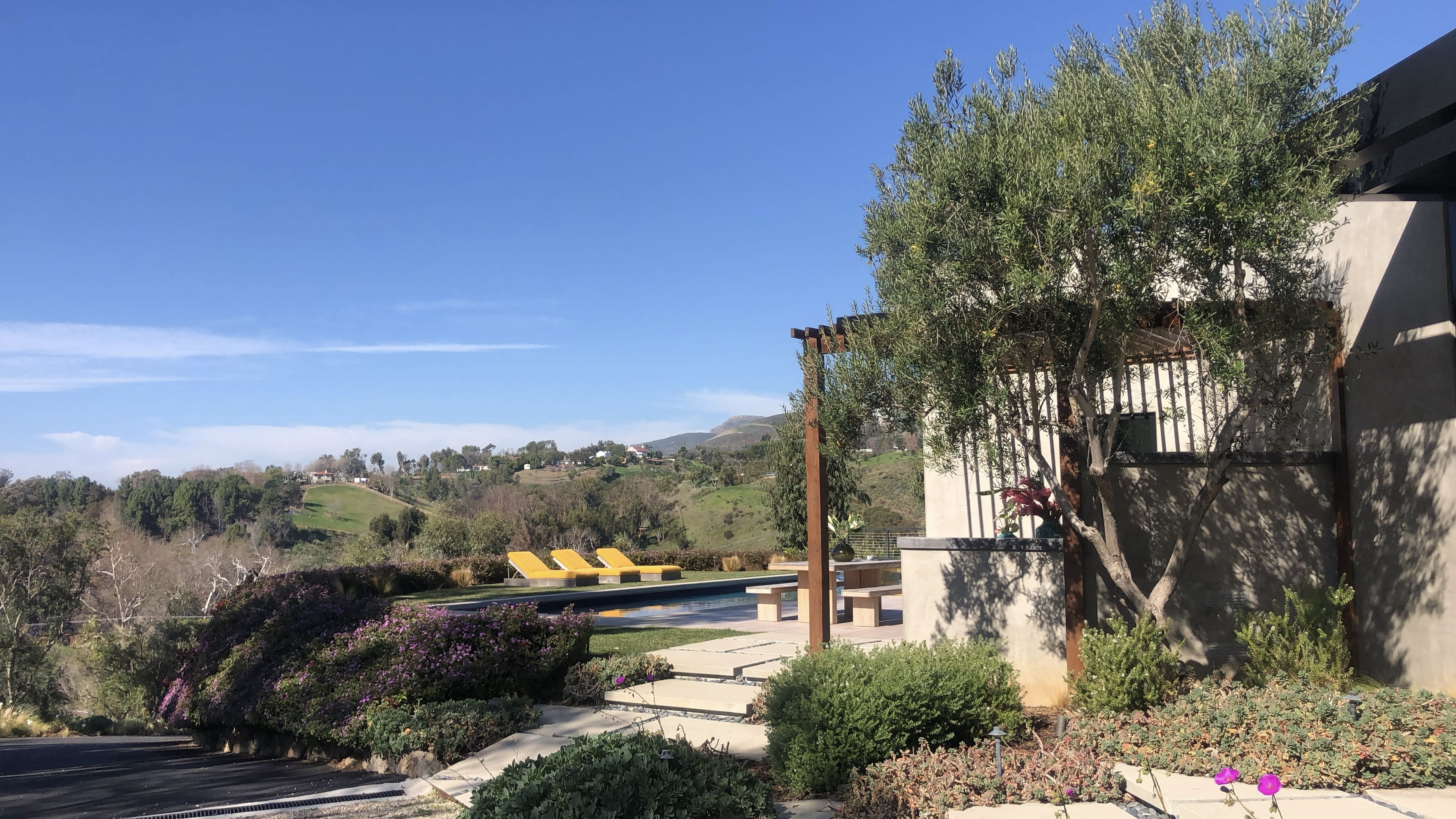 A modern house with a pool and lounging chairs is surrounded by landscaped gardens and hills under a clear blue sky.
