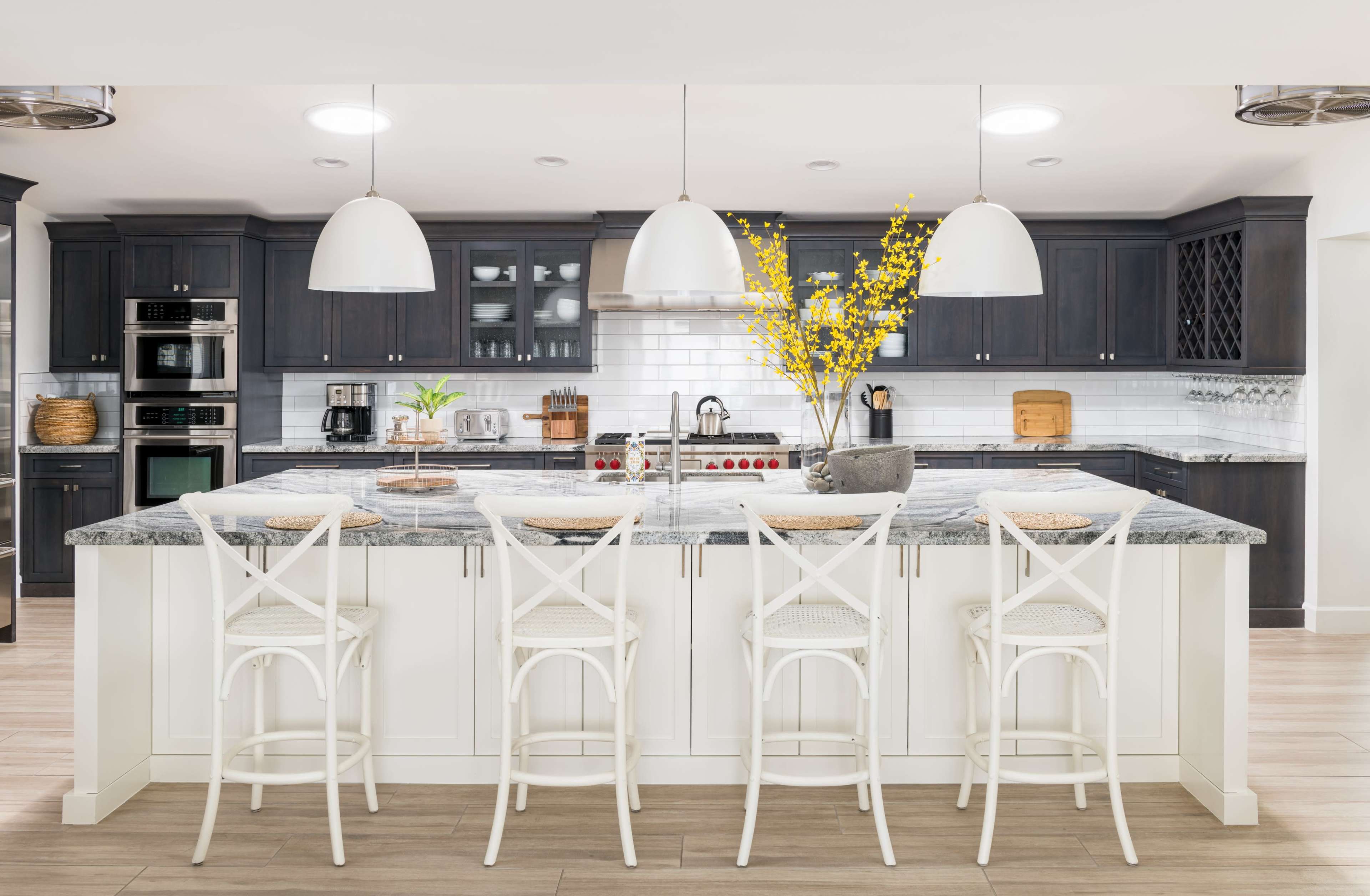 A modern kitchen features a large island with four white stools, dark cabinetry, stainless steel appliances, and decorative yellow flowers in a central vase.