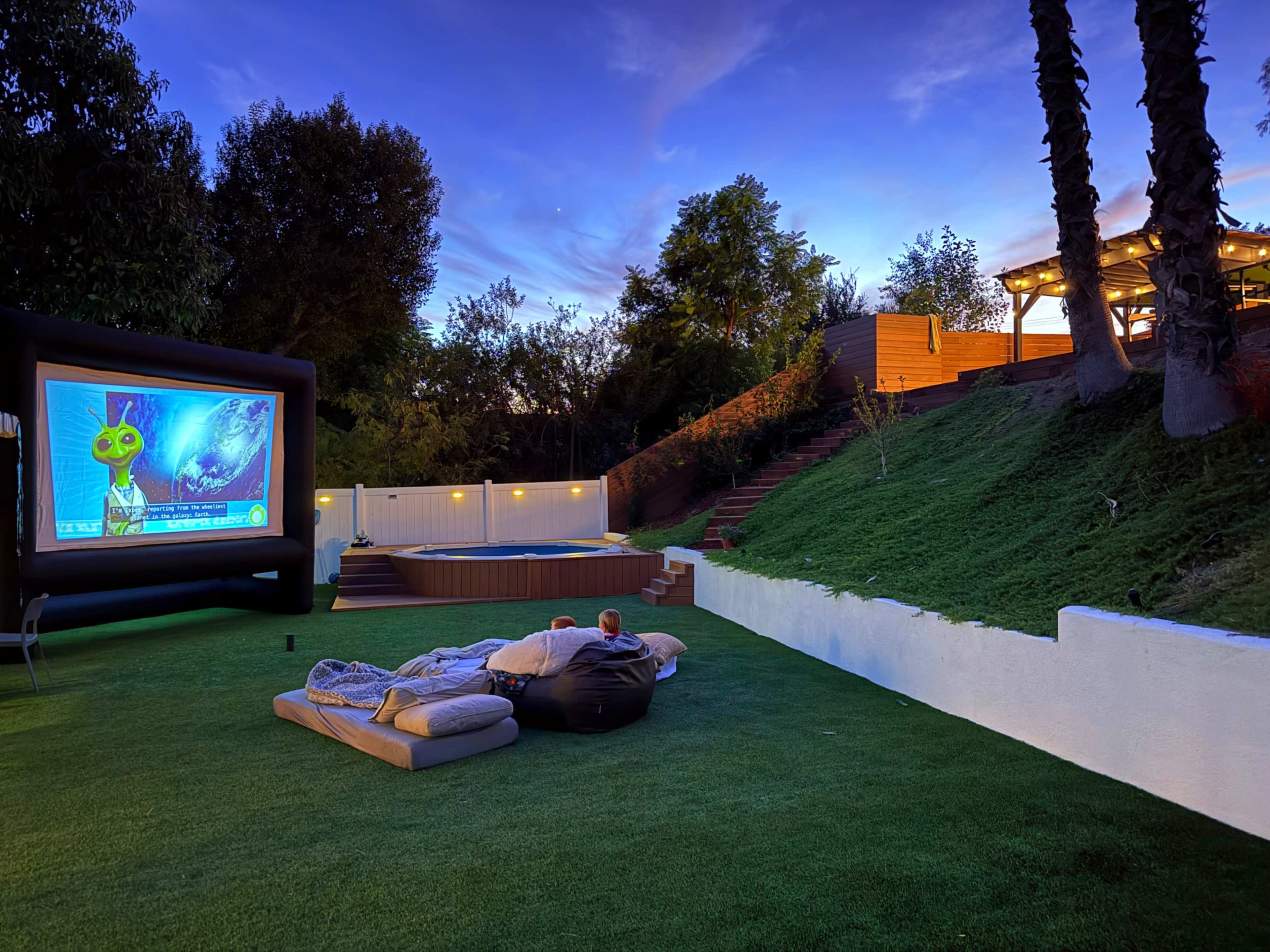 A backyard features an inflatable movie screen, comfortable seating on a grassy lawn, and a view of a hot tub and illuminated patio in the evening.