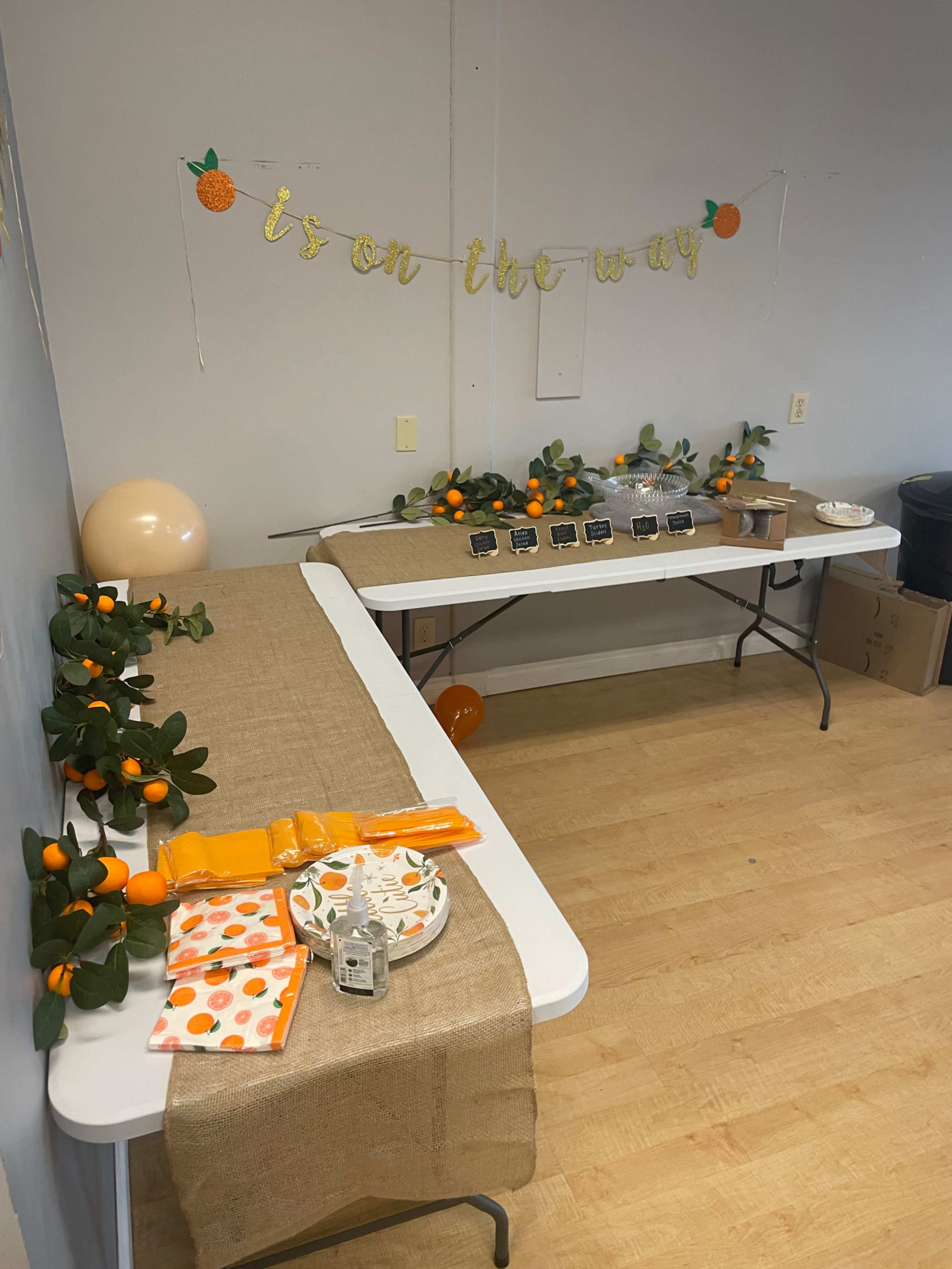 The scene shows a decorated room with two tables, one adorned with a burlap runner, small signs, and orange-themed decorations, while a banner reading "is on the way" hangs on the wall.
