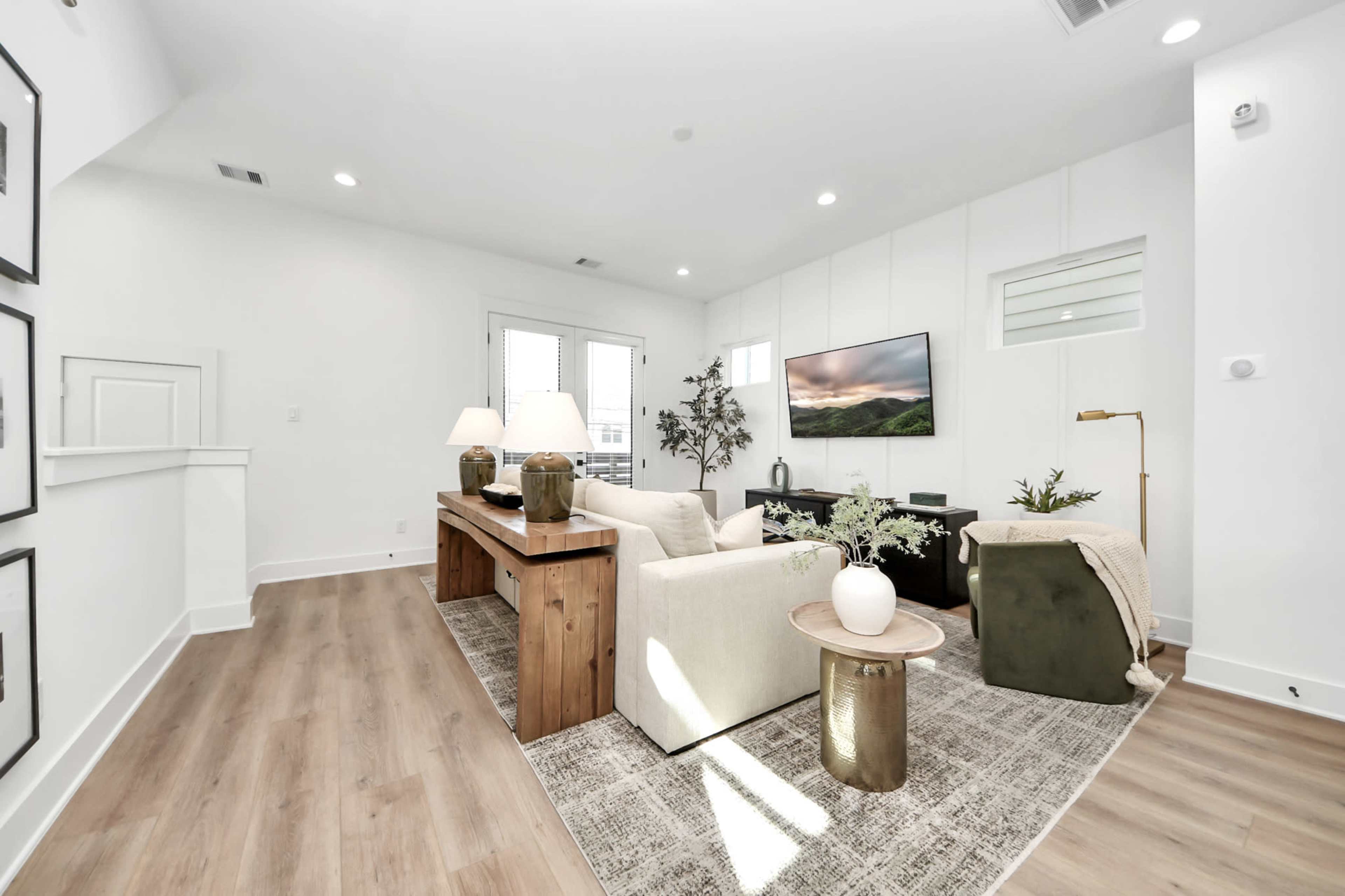 The image shows a bright living room with a large television mounted on the wall, a wooden table, and light-colored furniture arranged around a patterned area rug.
