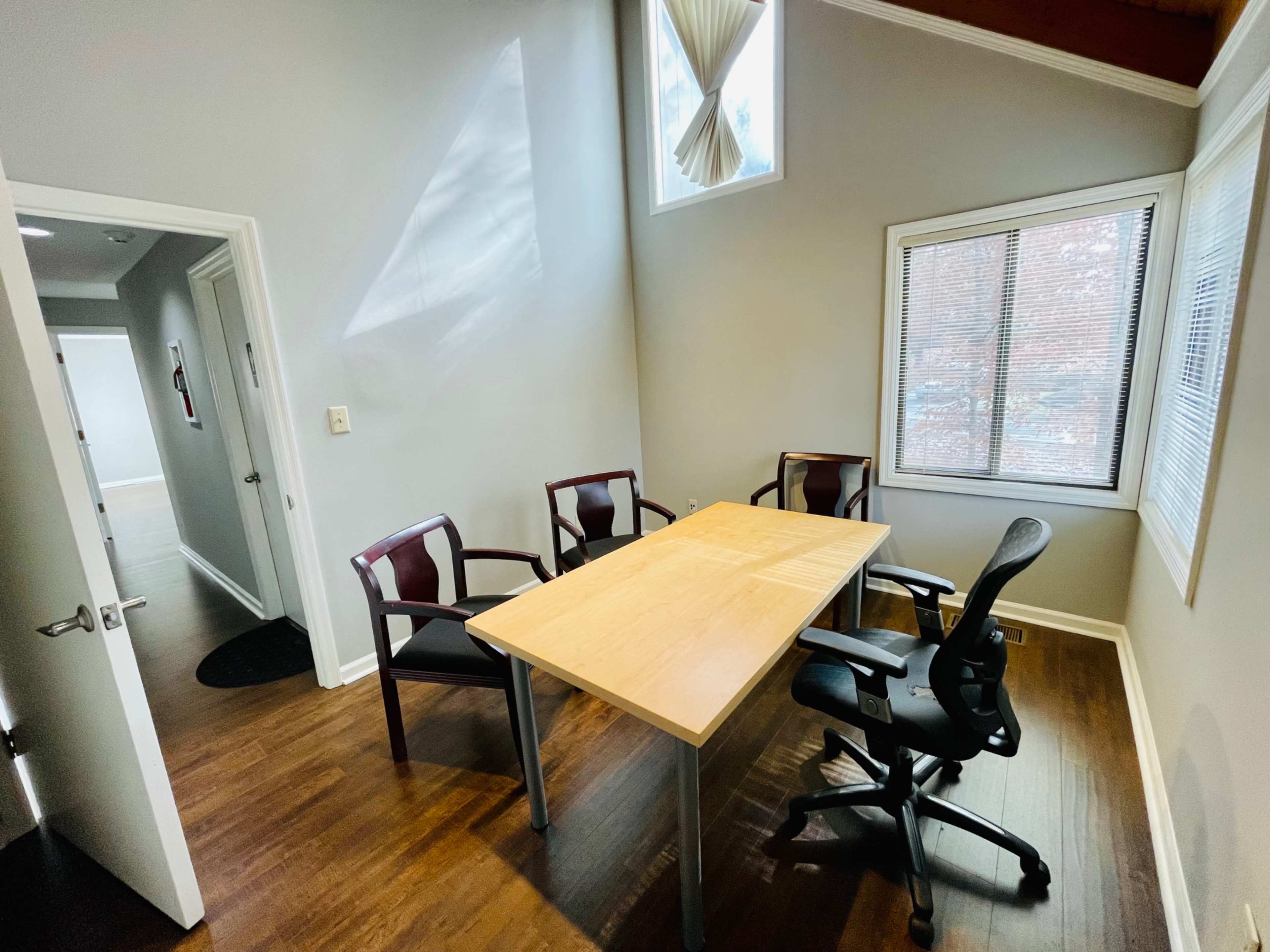 The image shows a small meeting room with a wooden table and four chairs, featuring a window with natural light.