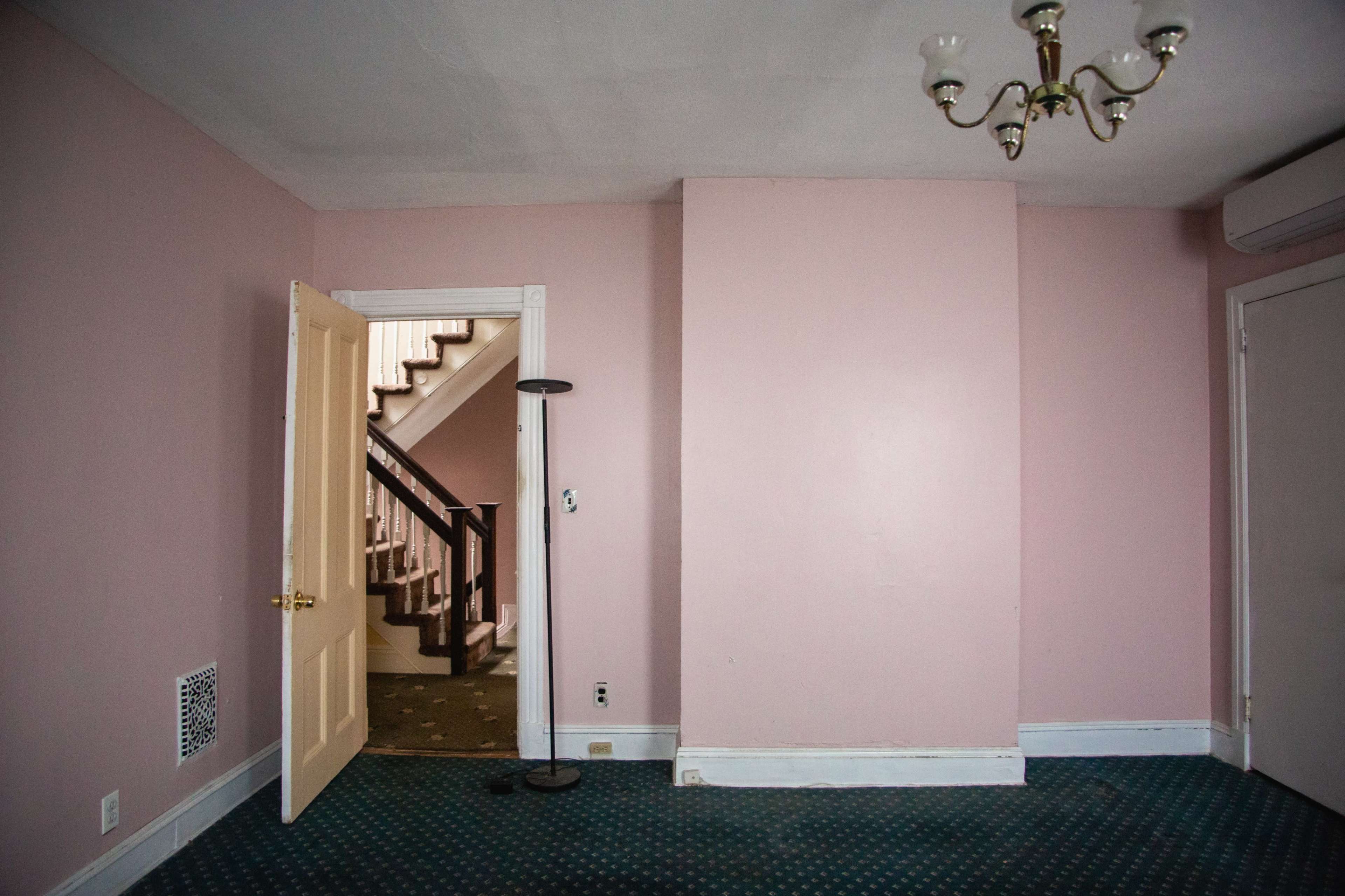 The image shows a sparsely furnished room with pink walls, a green carpet, an open door leading to a staircase, and a chandelier overhead.