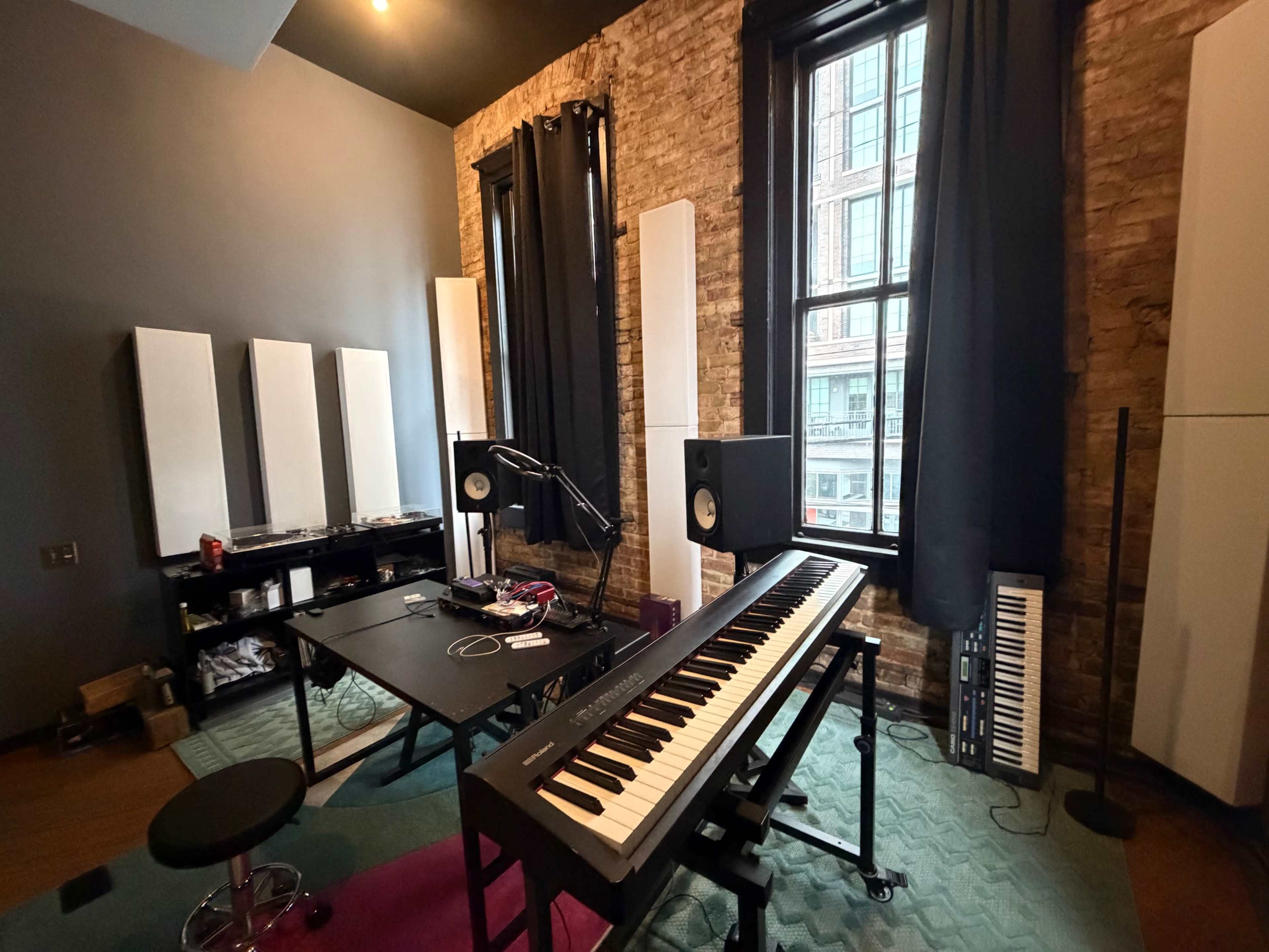 The image shows a music studio featuring a keyboard, audio monitors, and soundproofing panels against exposed brick walls, with large windows allowing natural light.