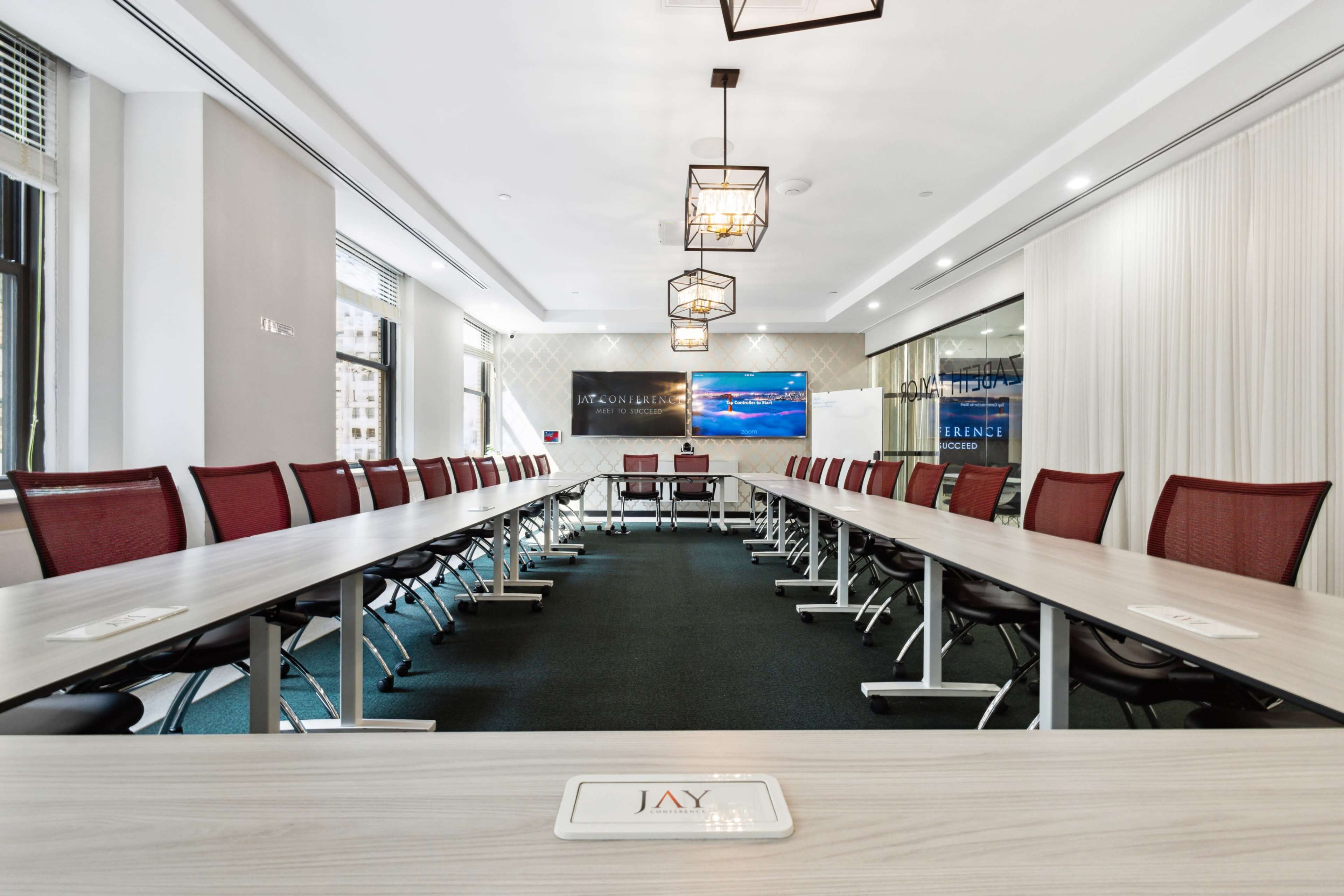 A spacious conference room with a long rectangular table, surrounding red chairs, and a presentation screen at one end.