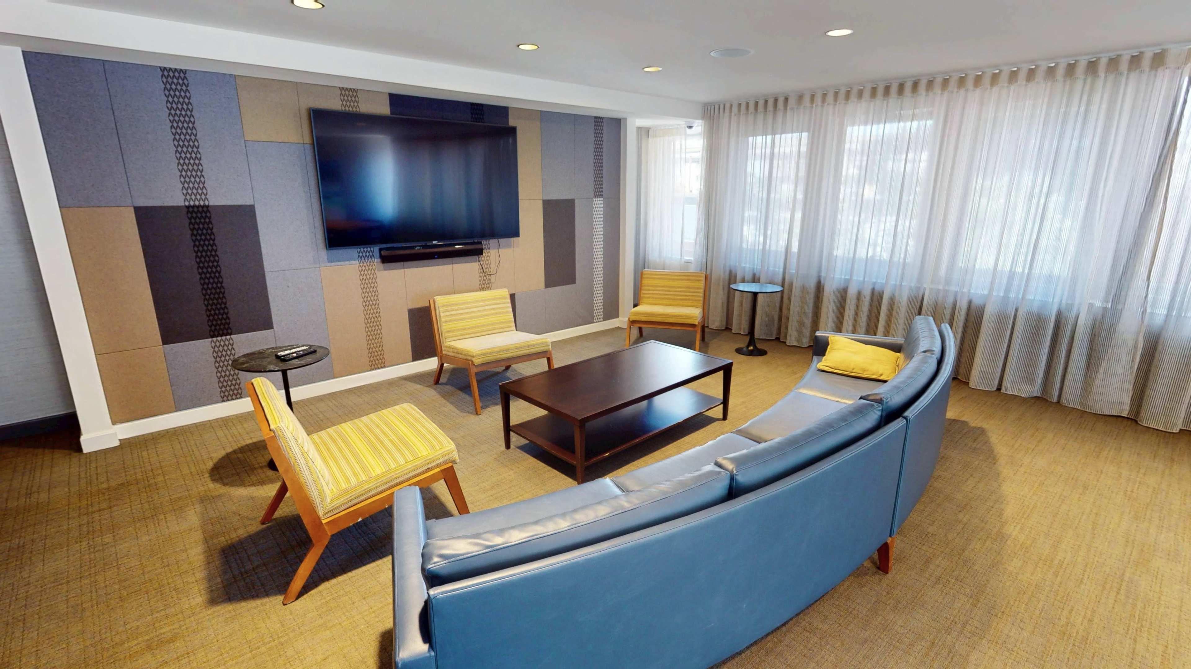 The image shows a modern living room with a large television mounted on the wall, a circular blue couch, two yellow-striped armchairs, and a coffee table in the center, surrounded by floor-to-ceiling windows with sheer curtains.