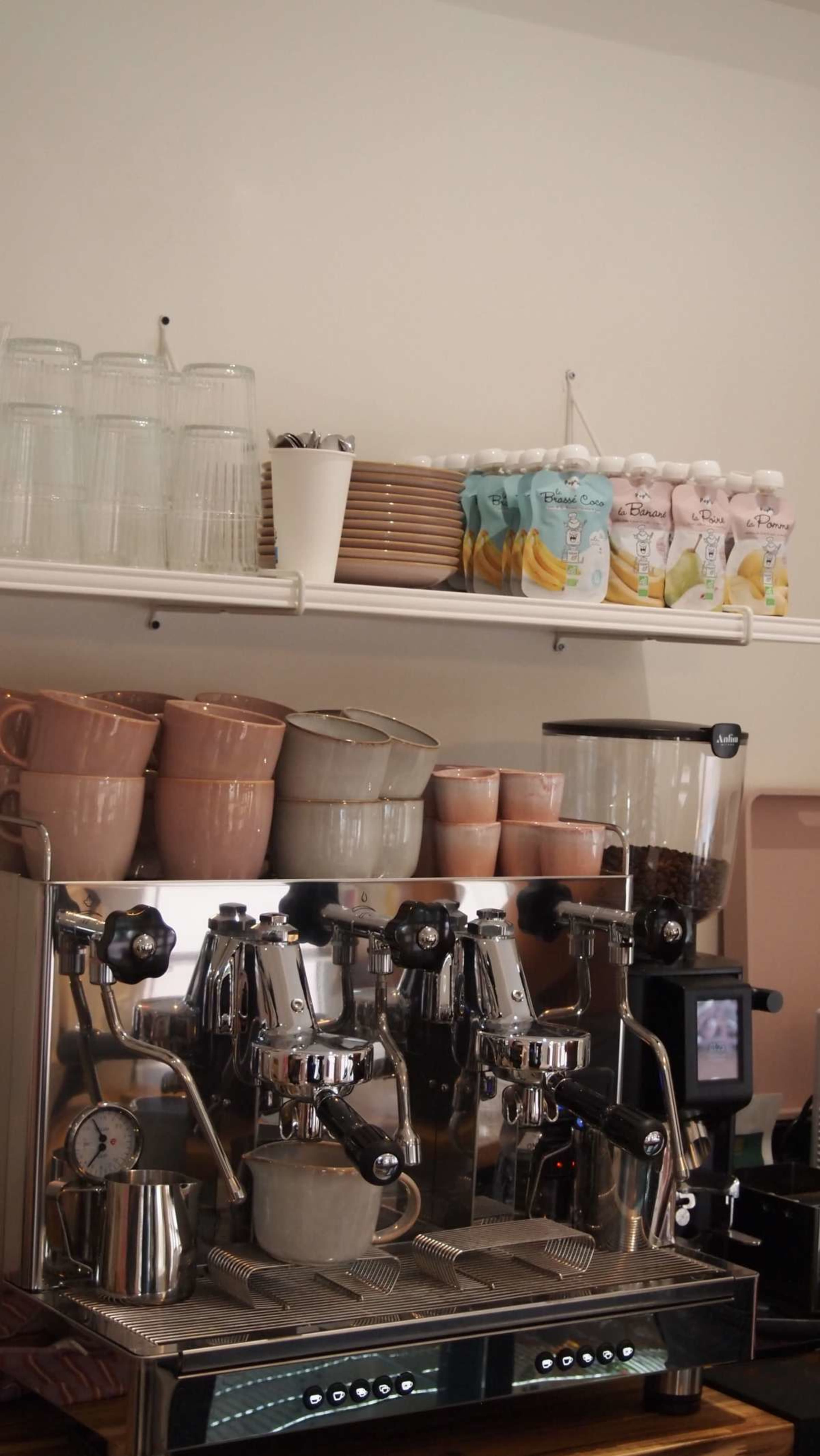 The image shows a coffee shop counter with an espresso machine, various mugs, and stacked cups on a shelf above.