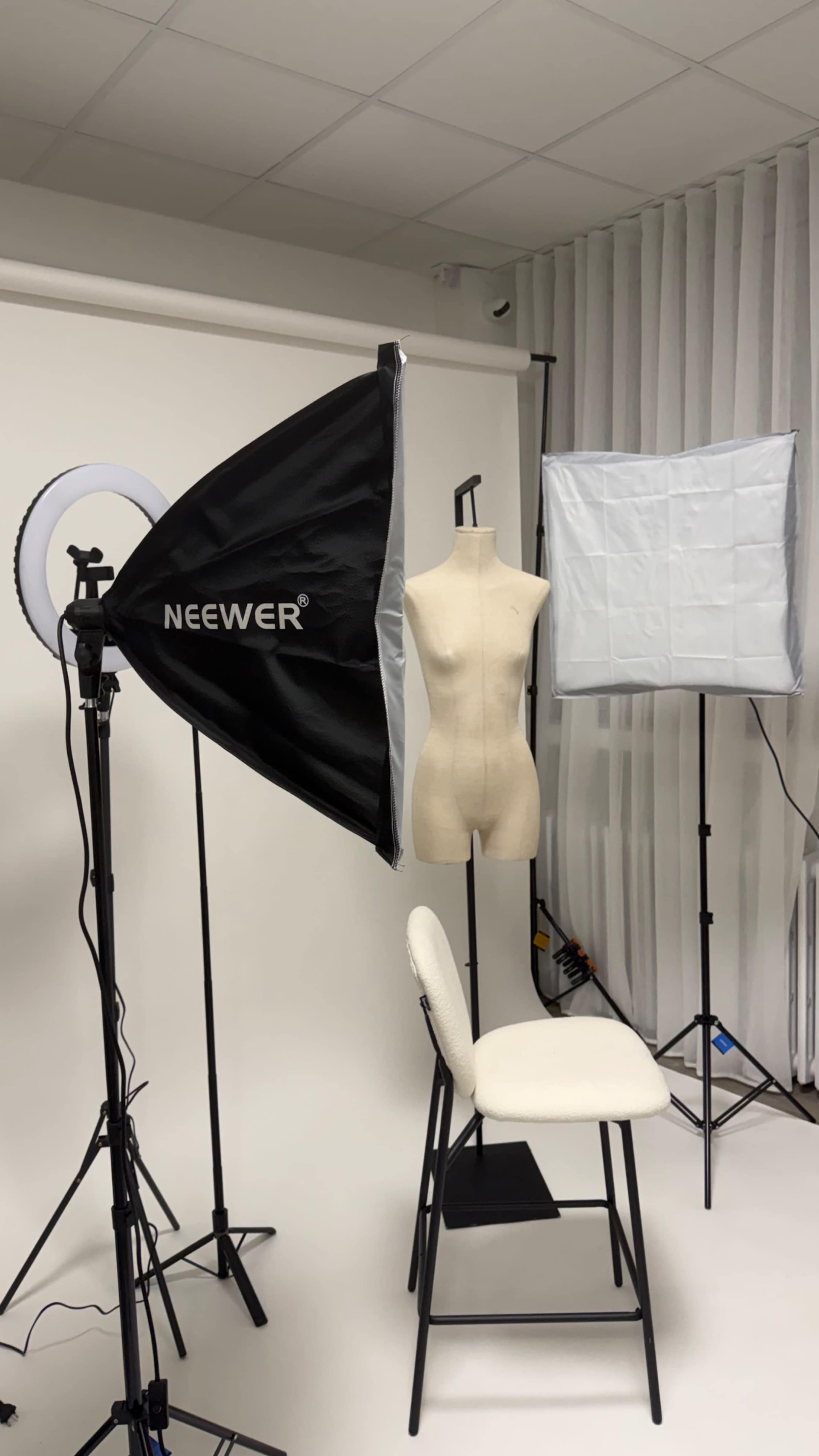 The image shows a photography studio setup with a mannequin on a chair, two softbox lights, and a ring light against a plain backdrop.