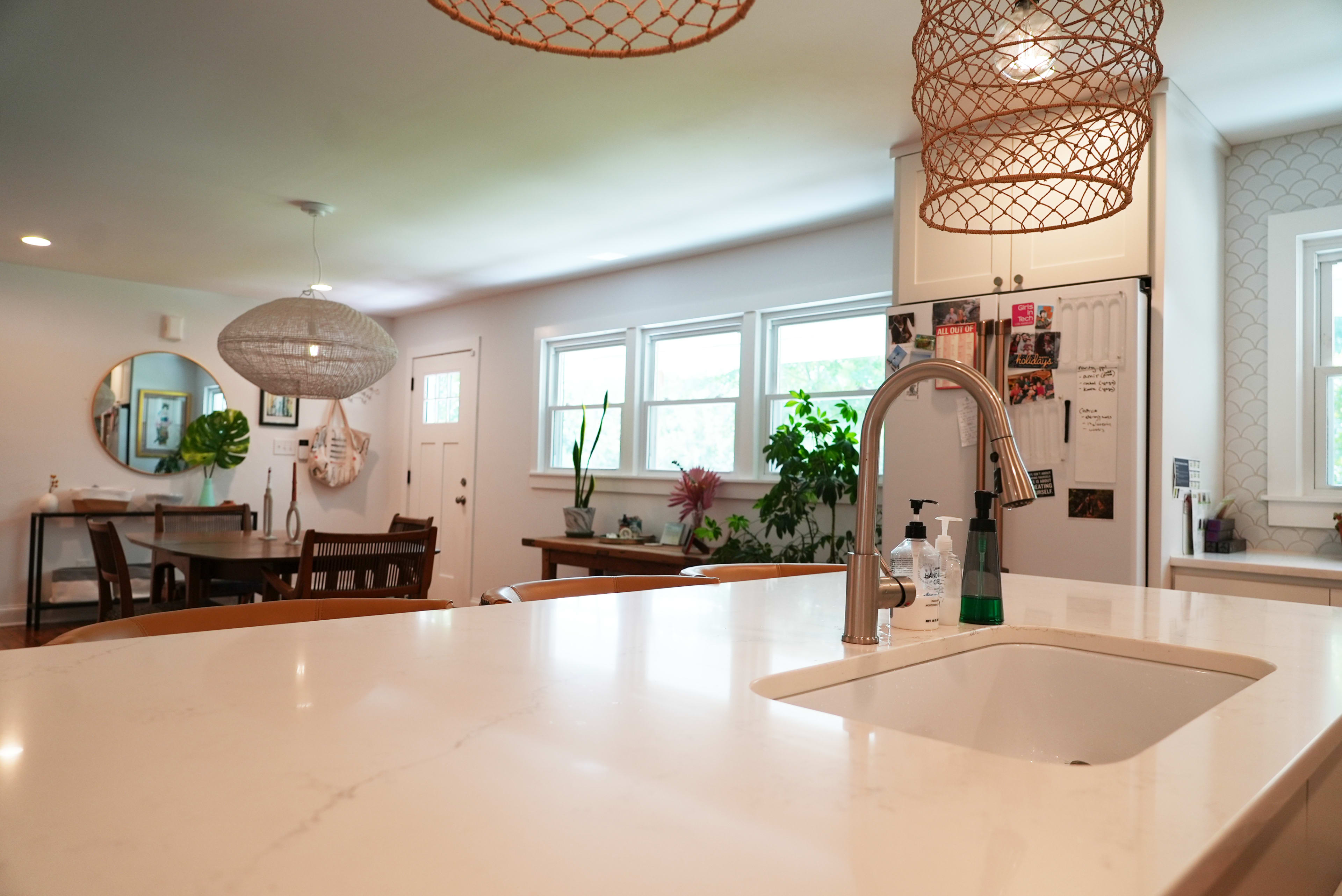 A modern kitchen features a large island with a sink, surrounded by woven pendant lights, and is connected to a dining area with a wooden table and various plants.
