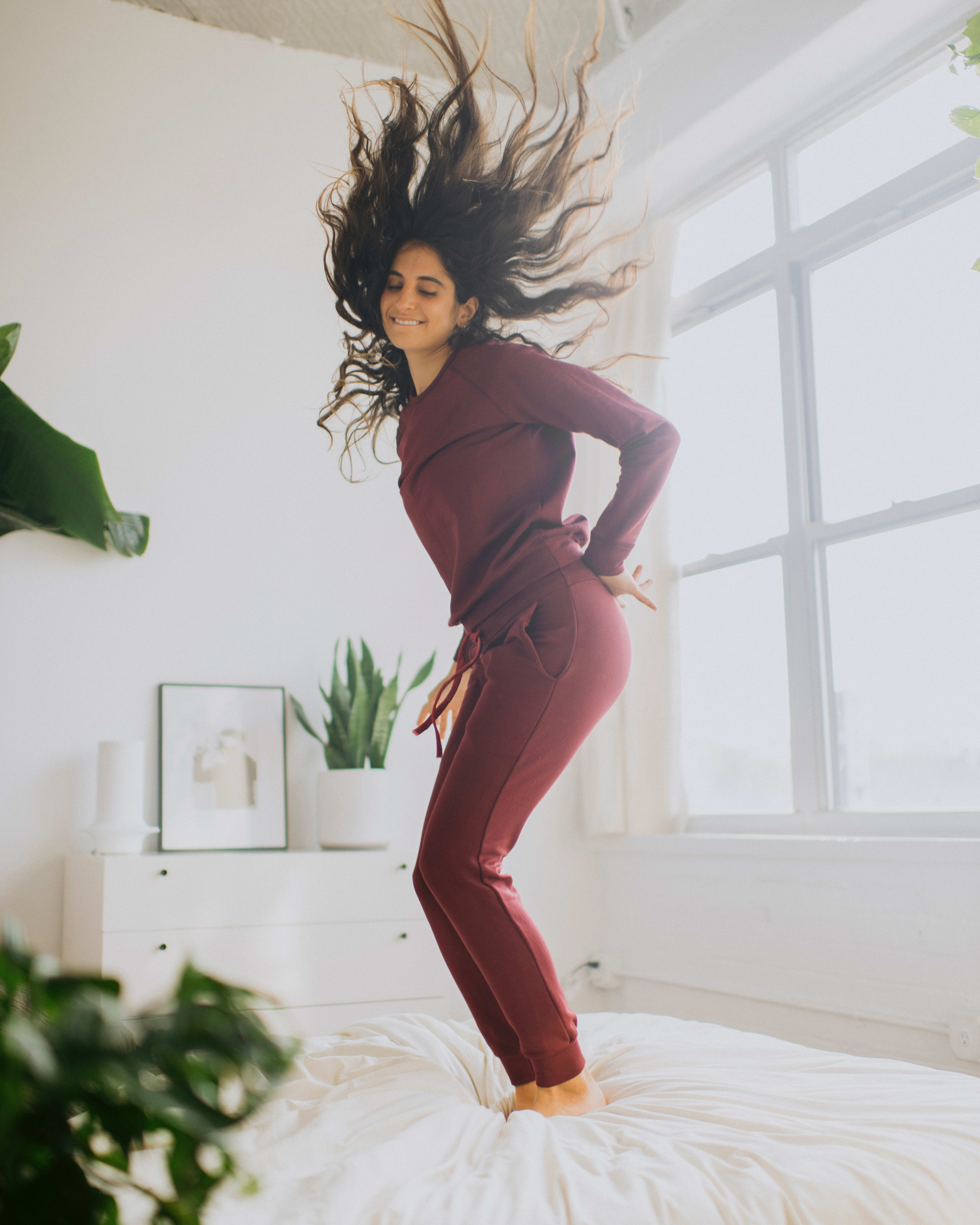A fashion photo shoot of a woman jumping on a white bed with her hair in the air.