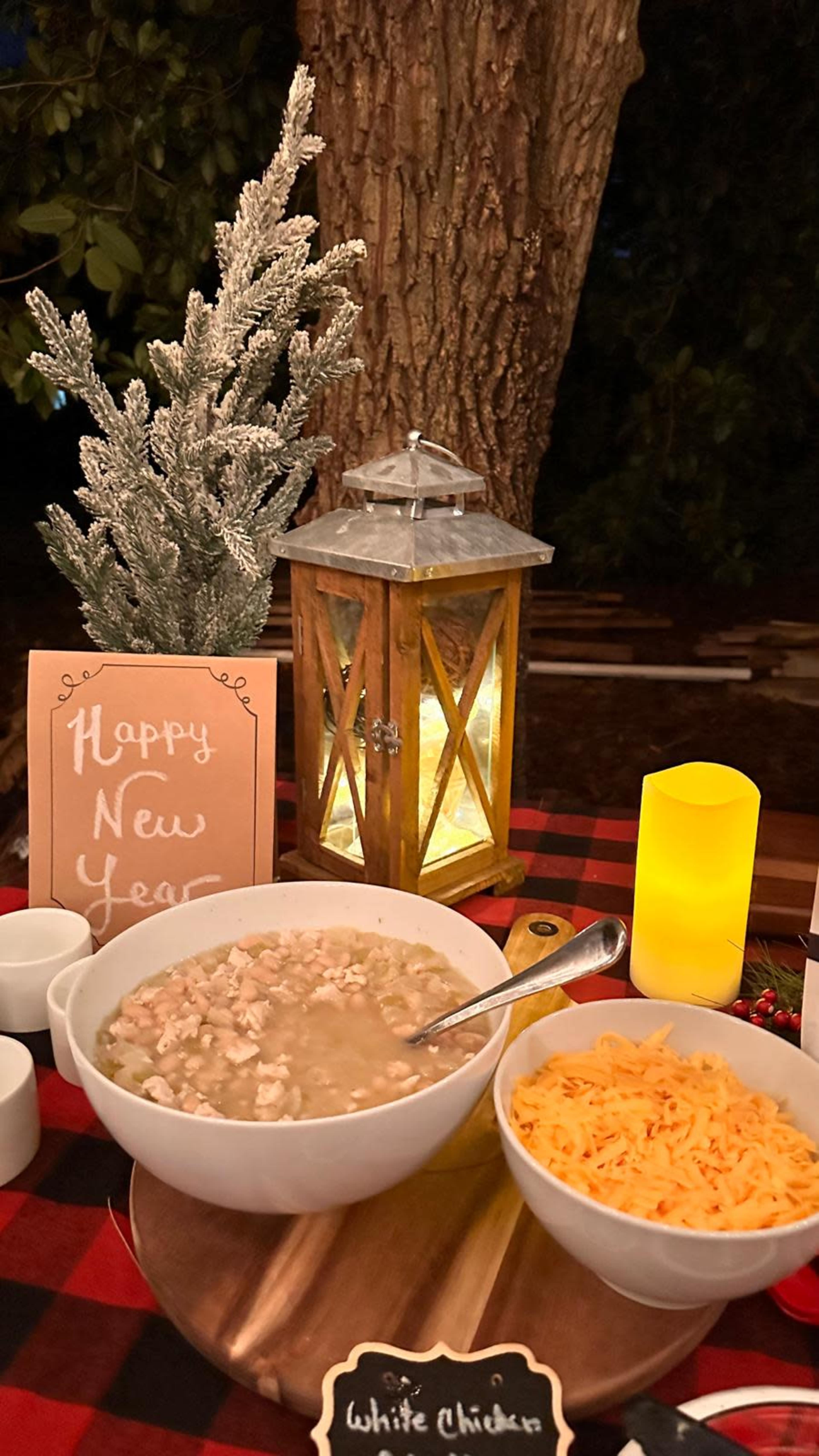 A table setting features a bowl of white chicken soup, a dish of shredded cheese, a decorative lantern, and a sign that reads "Happy New Year," all arranged with festive decorations.