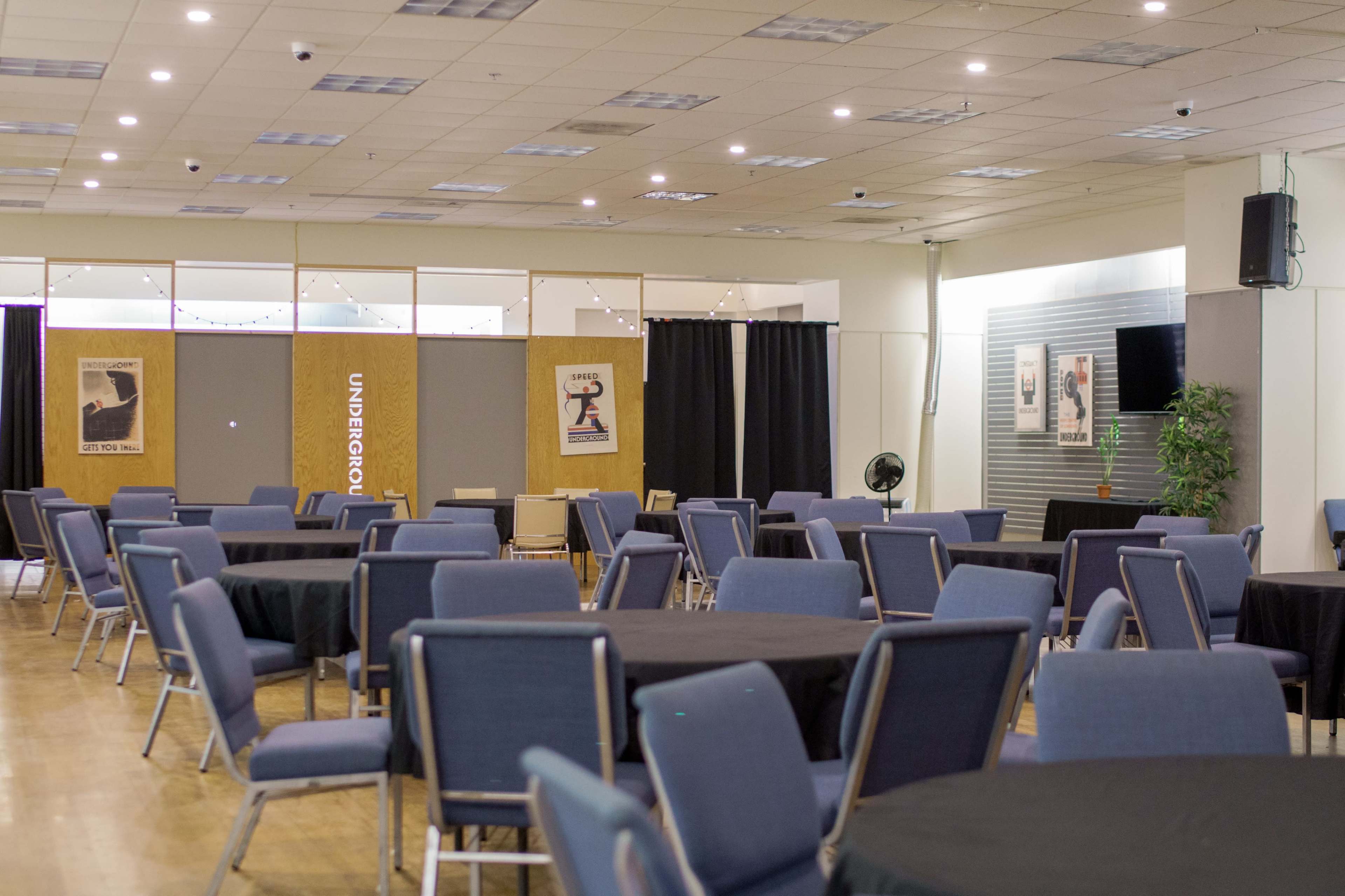 The image shows an empty indoor event space with several round tables and chairs arranged, along with promotional posters on the walls.