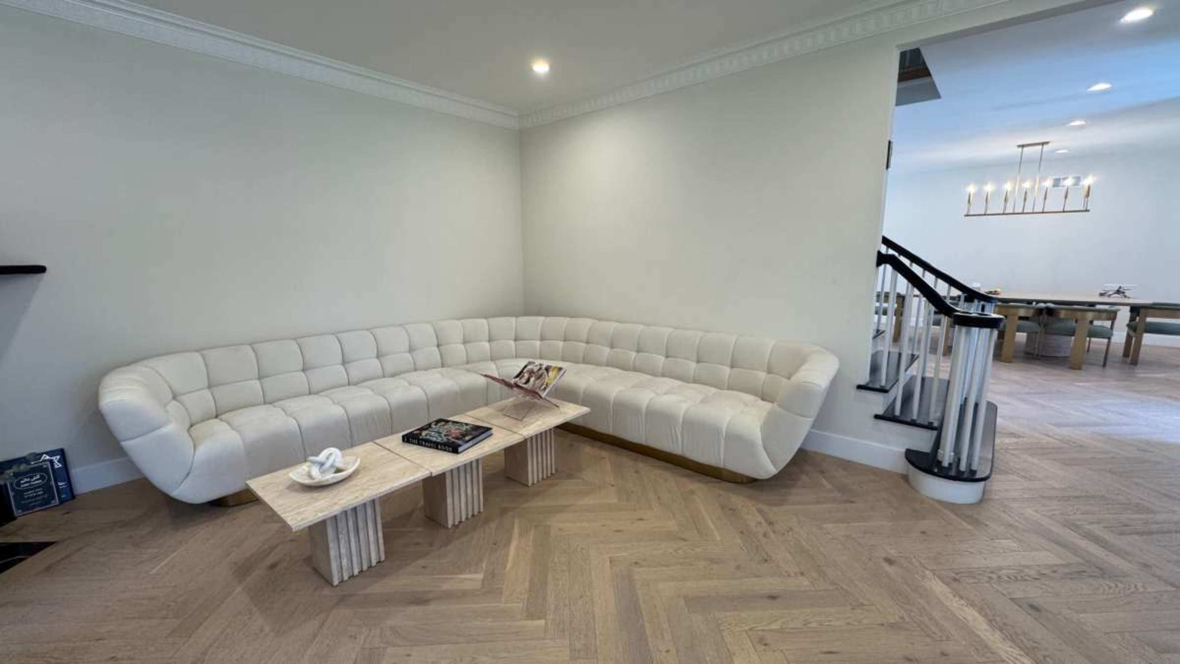 The image shows a modern living room with a curved white couch and a minimalist coffee table, featuring wood flooring and a staircase in the background.