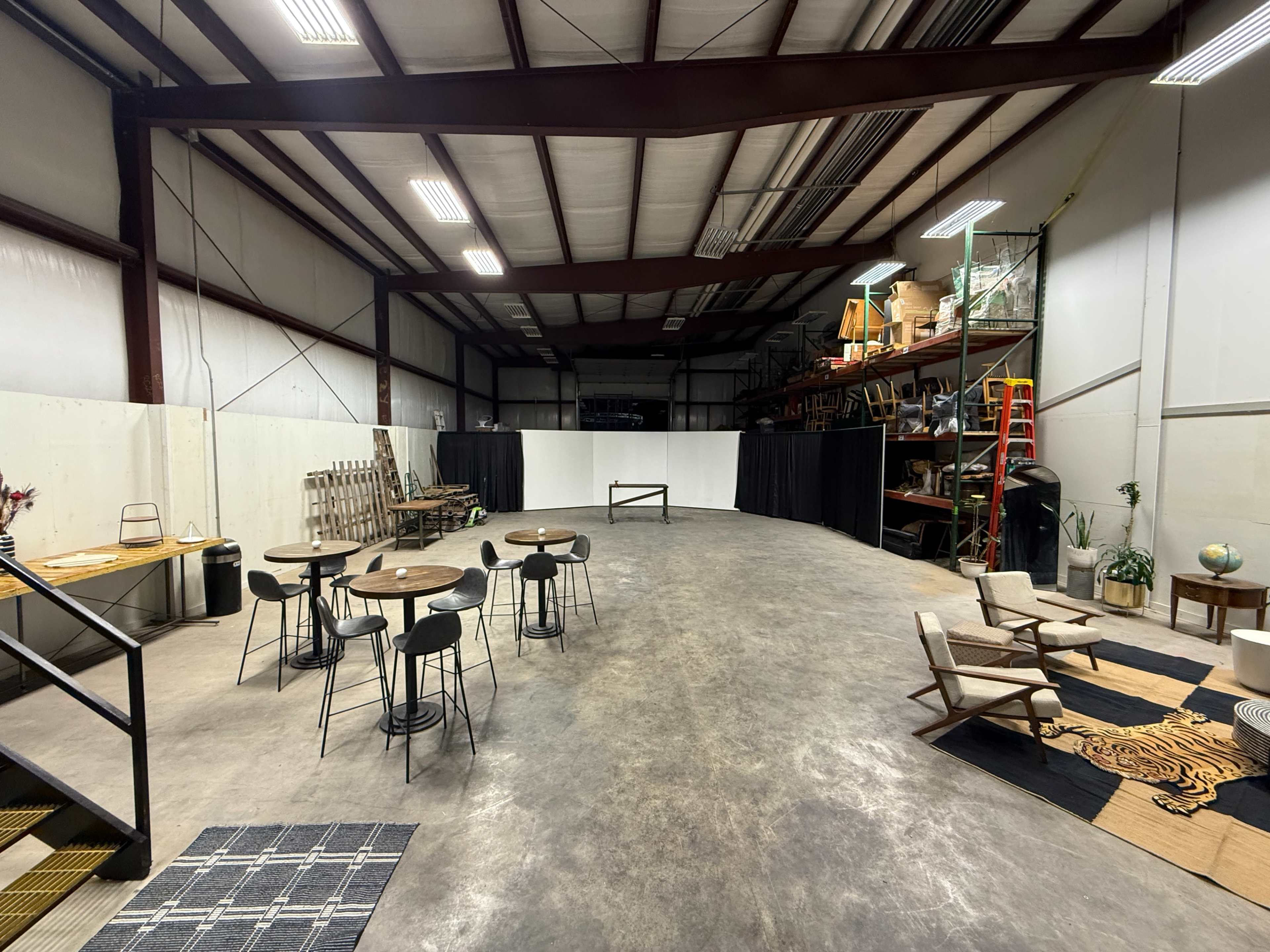 The image shows an open warehouse space with wooden flooring, tables and chairs set up in one area, and shelves containing various items in the background.