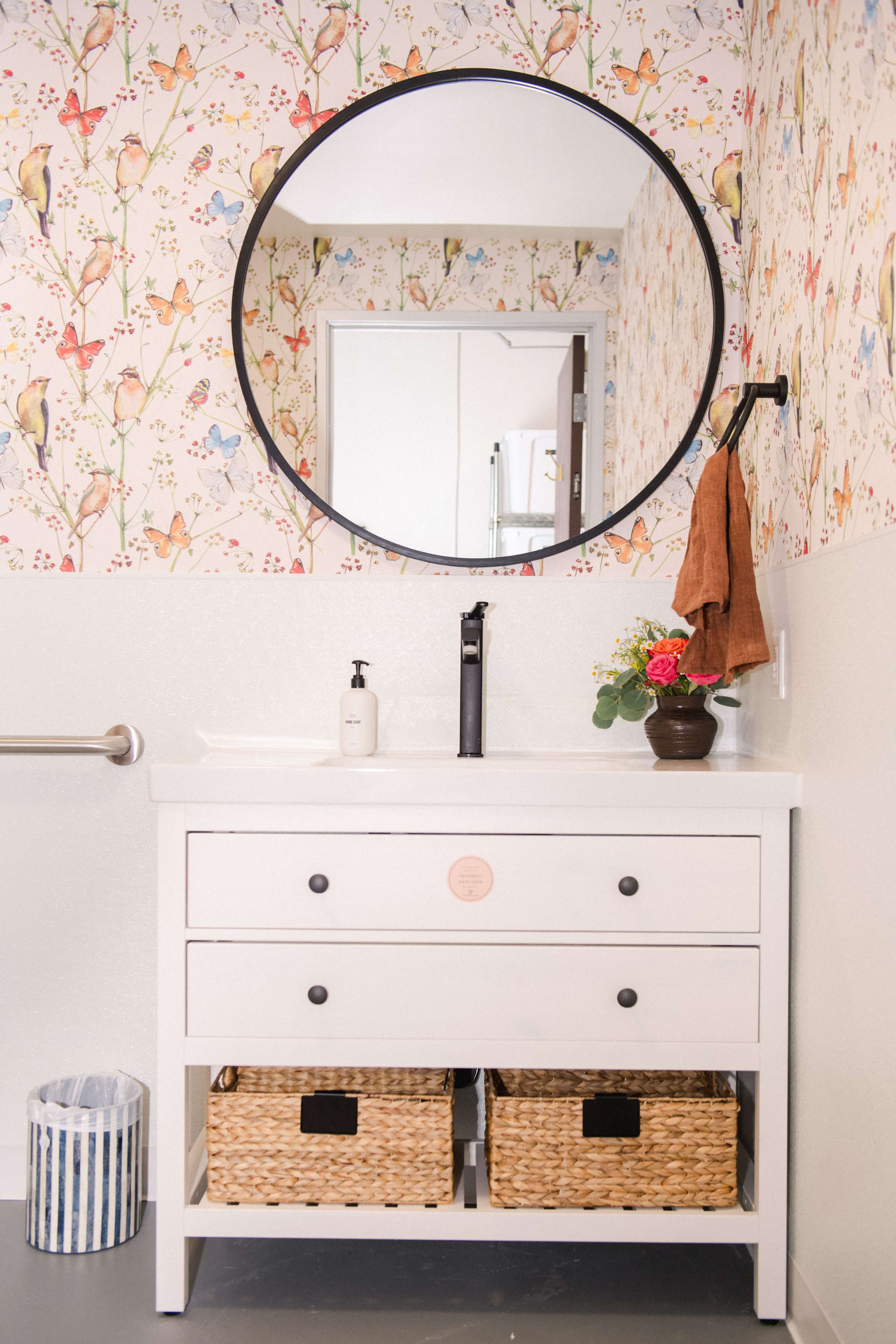 A bathroom with a round mirror above a white vanity featuring two drawers and woven baskets below, set against a colorful bird-themed wallpaper.