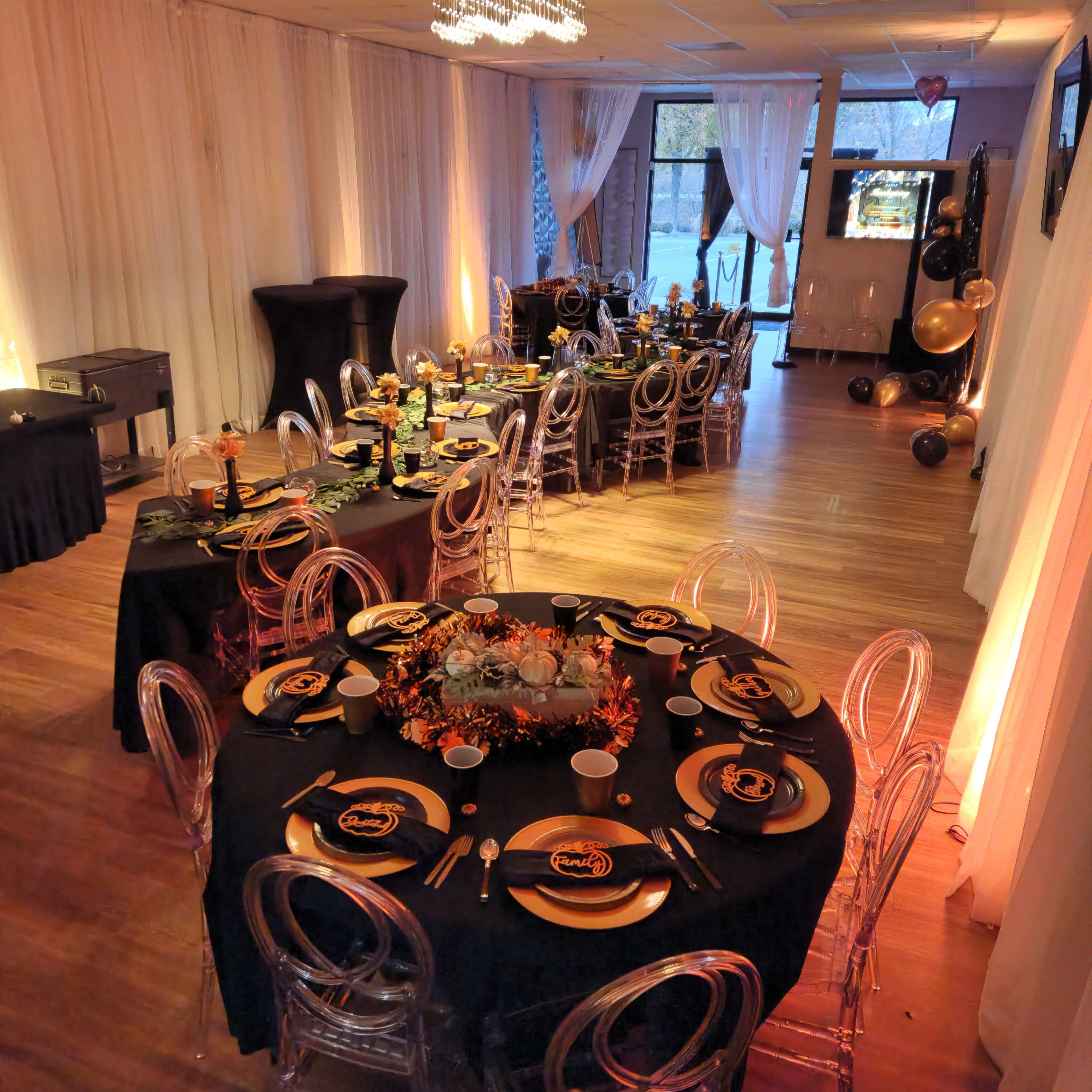 A decorated event space features tables set with black tablecloths, gold accents, and clear chairs, with a backdrop of balloons and a viewing screen.