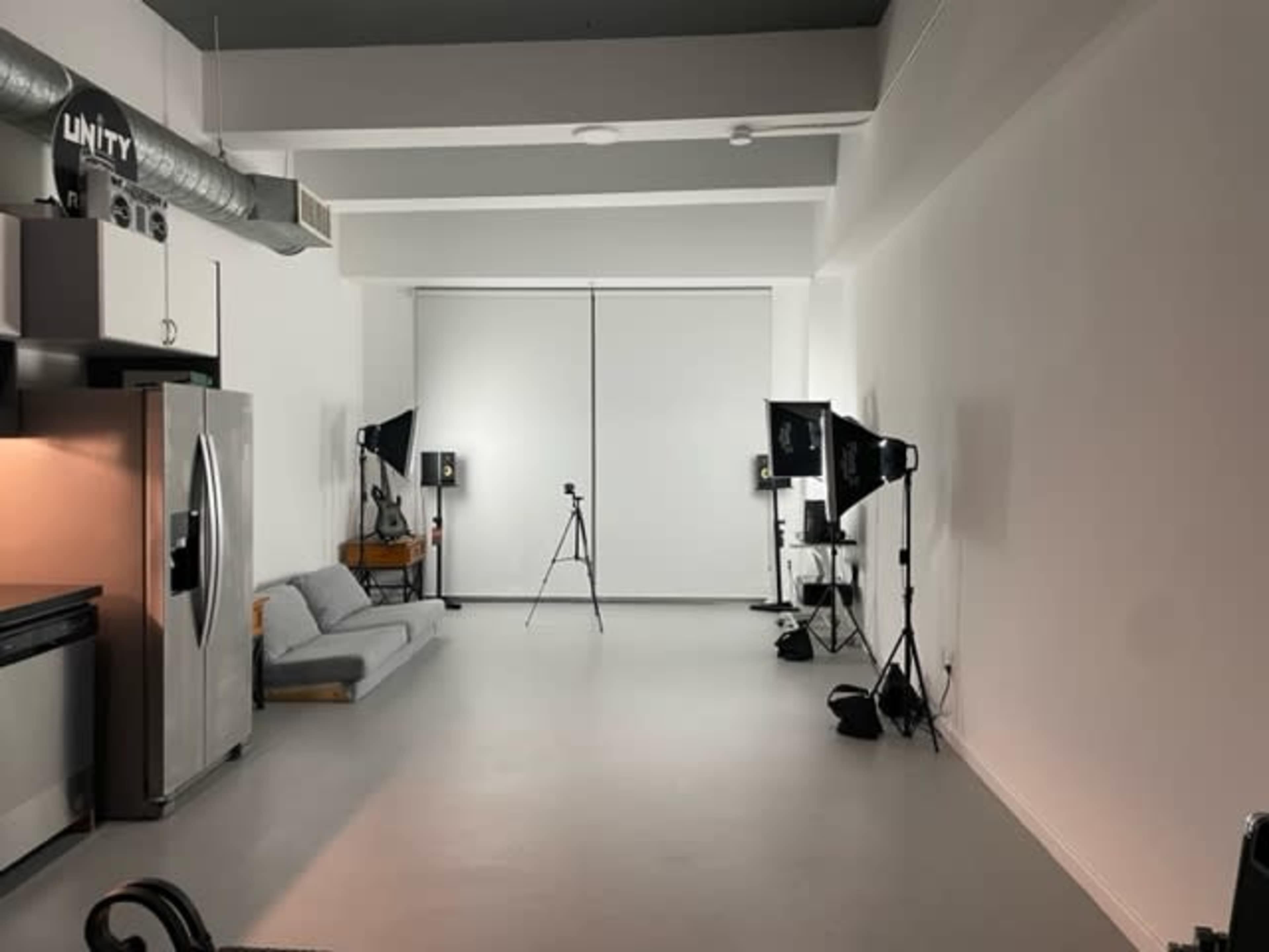 A spacious studio is equipped with a gray sofa, a kitchen area, and photography lighting setups on either side.