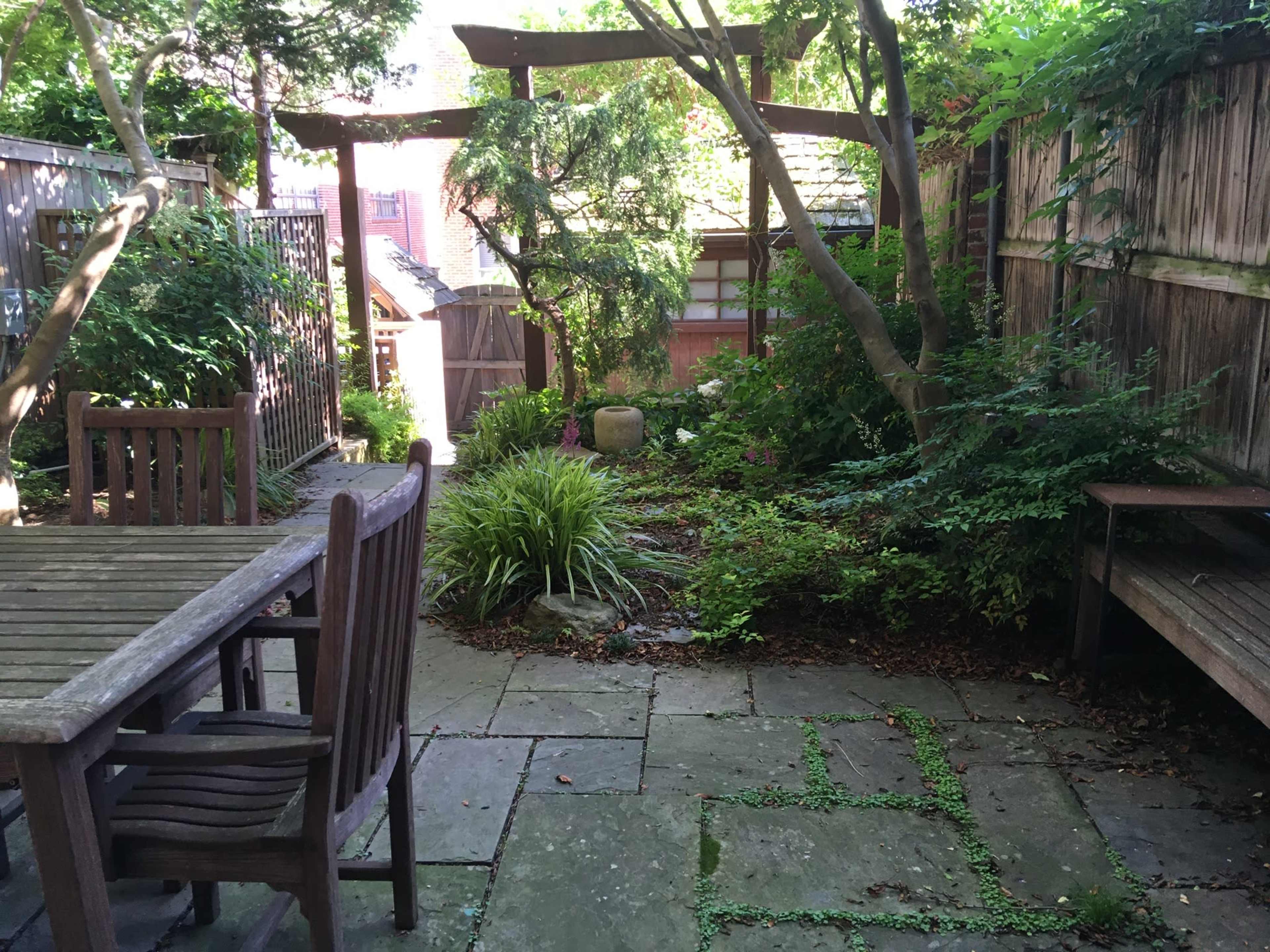 A stone-paved garden area with wooden furniture, surrounded by greenery and a trellis overhead.