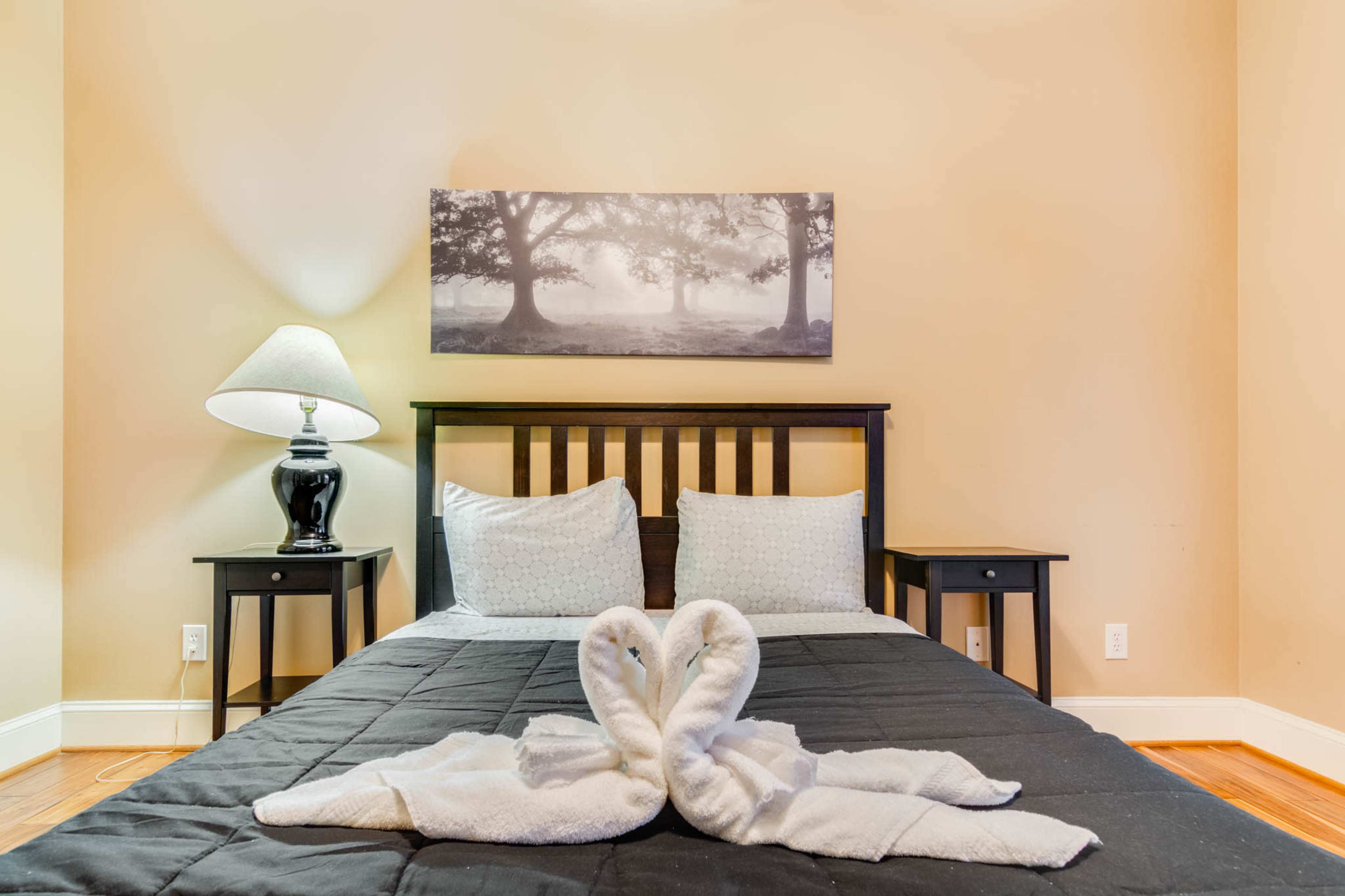 The image shows a simple bedroom with a bed, a black lamp, and a decorative wall art featuring trees.