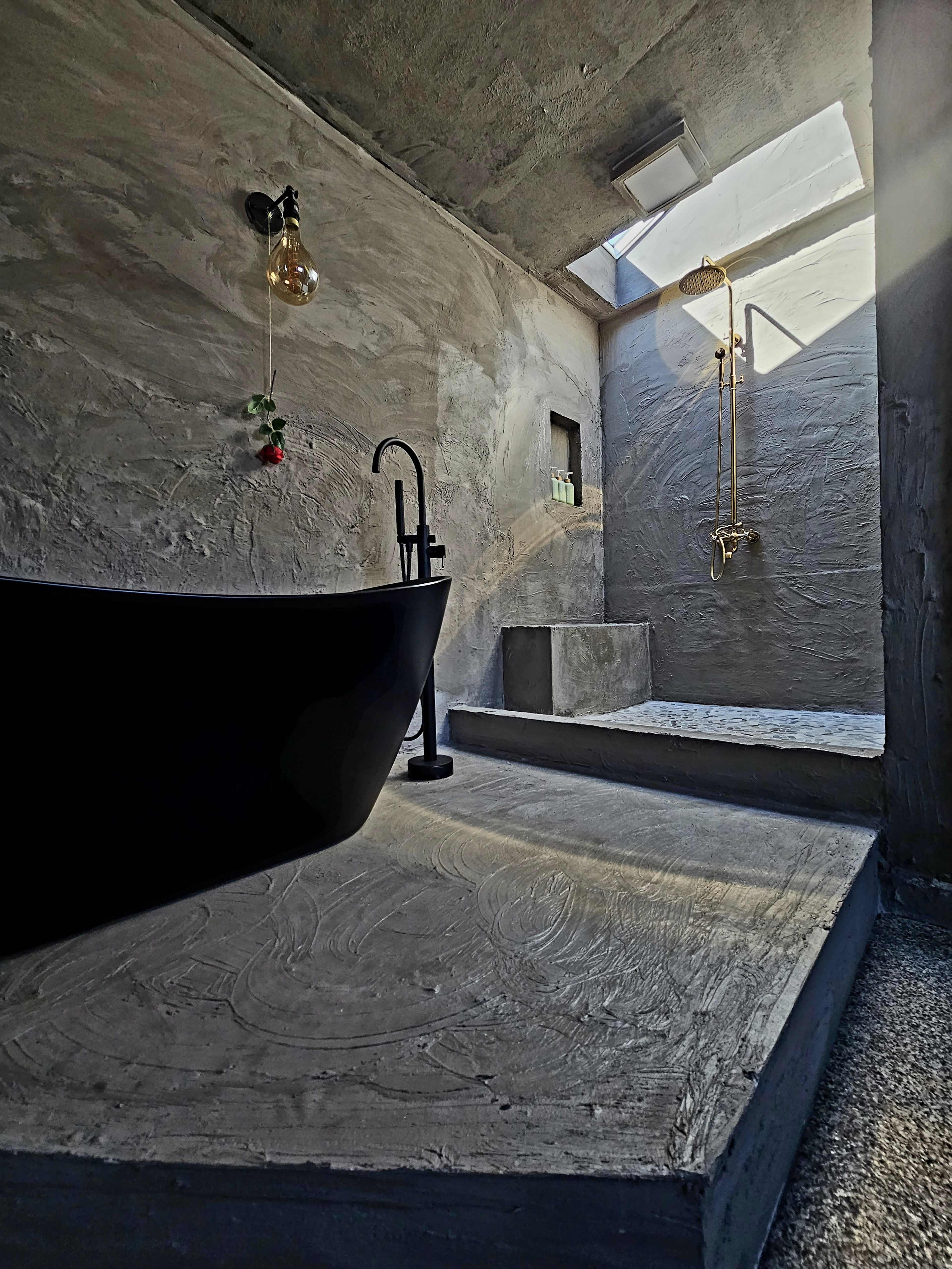 The image shows a minimalist bathroom featuring a black freestanding bathtub, a gold wall-mounted shower fixture, and a skylight above a textured concrete floor.