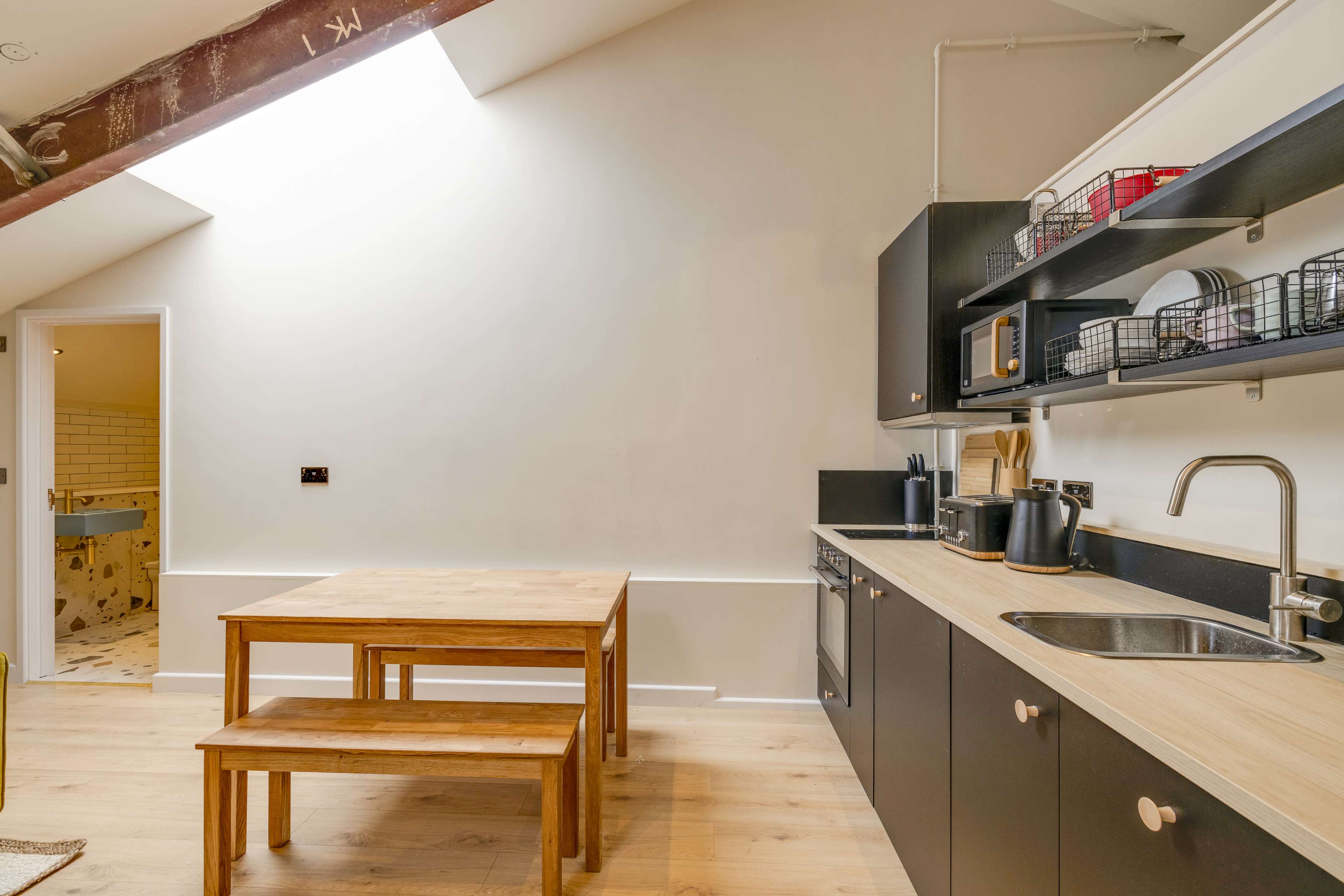 A modern kitchen featuring a wooden table with benches, dark cabinetry, and an open layout illuminated by a skylight.
